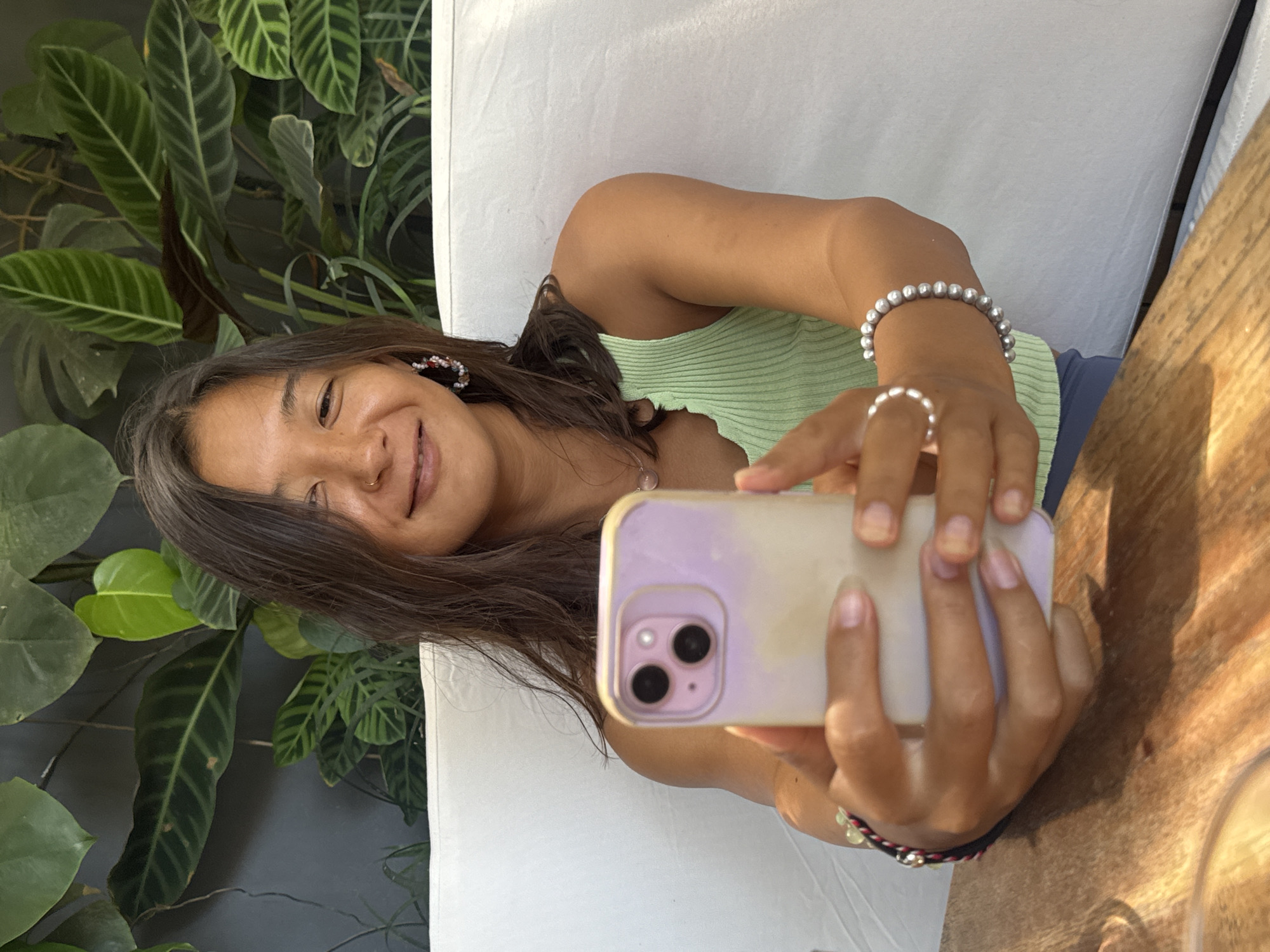 A young woman is captured in a vertical selfie, smiling warmly at the camera. She has long, dark hair, and wears a light green ribbed tank top. Her accessories include beaded hoop earrings, a nose ring, a delicate necklace with a light pink pendant, a pearl bracelet on her right wrist, and a pearl ring on her right index finger. She holds a smartphone with a light purple/lavender case in her hands, taking the picture herself. The phone's two dark camera lenses are visible on its back.
The setting appears to be an outdoor or semi-outdoor space, possibly a cafe or restaurant in Kuta Selatan, Indonesia. Behind her, a lush wall of various tropical green plants with prominent leaves, including species resembling Calathea and Monstera, creates a vibrant backdrop. A white cushioned surface is visible directly behind her, suggesting a chair or bench. To her right, a warm-toned wooden table surface, possibly dappled with sunlight, occupies the foreground, with part of a glass or cup also visible.
The bright, natural lighting indicates it is daytime, likely a sunny morning or afternoon, casting gentle light on her face and the surrounding elements. Her joyful expression conveys a relaxed and happy mood, fitting for the tropical and leisurely environment of Kuta Selatan. There is no visible text in the image.