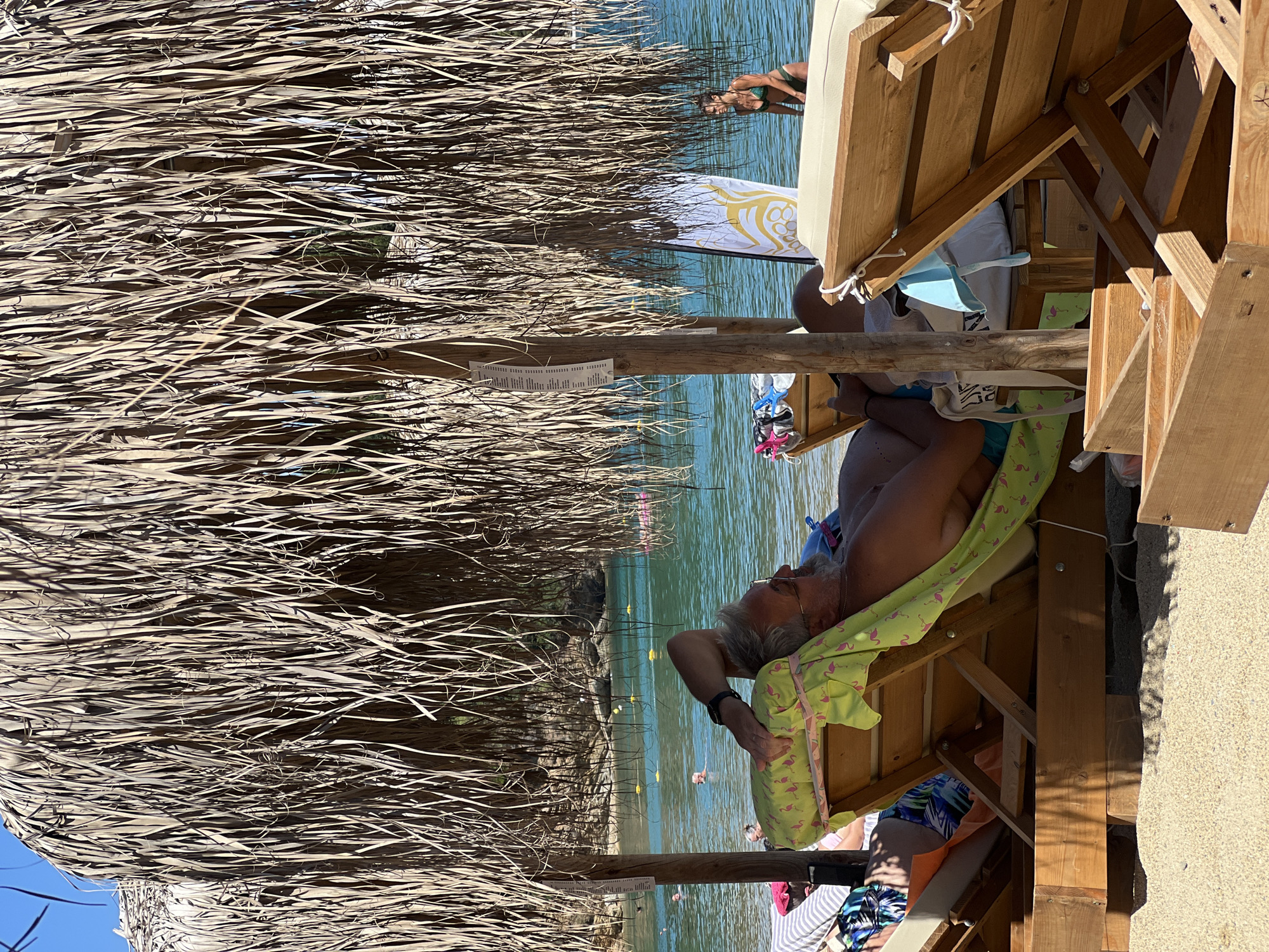 The image depicts a beach scene in Achladias, Greece, under a thatched umbrella. A man with graying hair is reclined on a sun lounger, wearing sunglasses and a chain. He is covered with a lime-green towel adorned with pink flamingos. In the background, the turquoise waters of the Aegean Sea are visible, with several people swimming and floating. A woman in a green bikini is standing near the water's edge, beside a surfboard. Wooden beach loungers and tables are scattered across the sandy shore. The overall atmosphere suggests a relaxed, sunny day at the beach.