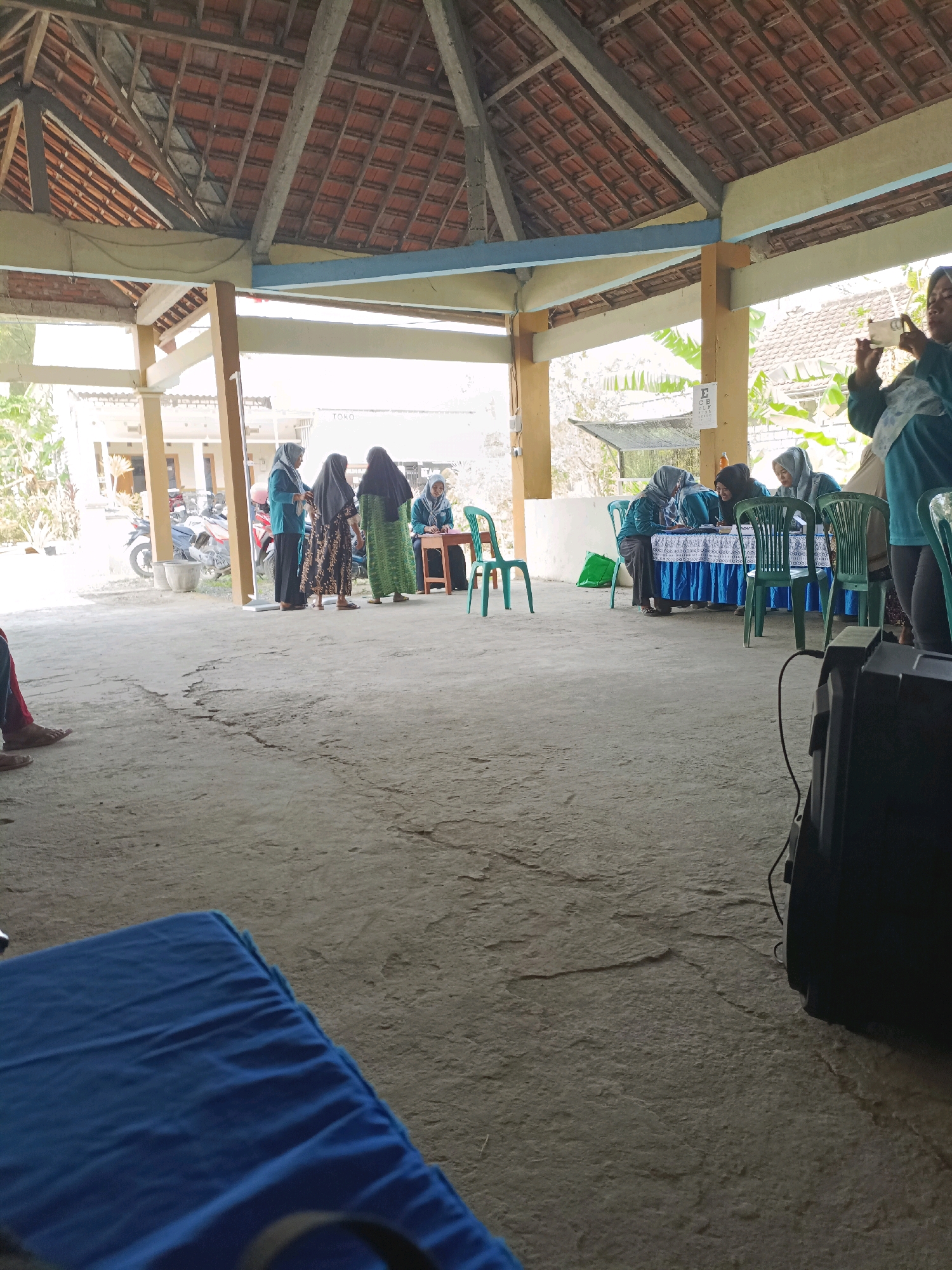 In Rancang Kencono, Indonesia, an outdoor gathering is taking place beneath a covered structure with a tiled roof. A group of women are seated at a table covered with a blue and white patterned cloth, engaged in an activity that appears to be administrative or educational. Several green chairs surround the table. To the left, another group of women are standing, some in conversation. In the background, a building and parked motorcycles are visible. A black speaker with a wire is positioned in the foreground, to the right. The ground is a rough concrete surface. A blue tarp is spread out in the lower left portion of the image.