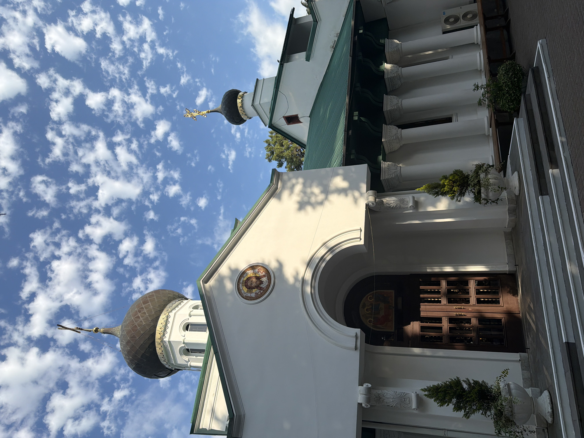 The image captures a view of the Nativity of the Theotokos Church in Sochi, Russia. The foreground features a white facade with a prominent arched entrance. Above the arch, a circular mosaic depicts the face of Christ. To the left and right of the entrance, potted evergreen trees flank a set of white stairs. The church has a distinctive green roof, supported by a colonnade of white pillars. Two domes are visible, one larger and darker, topped with a cross, and a smaller, darker dome also crowned with a cross. The sky is a vibrant blue, scattered with fluffy white clouds. The lighting suggests it is either early morning or late afternoon, with shadows cast across the scene. No people are visible in the image.