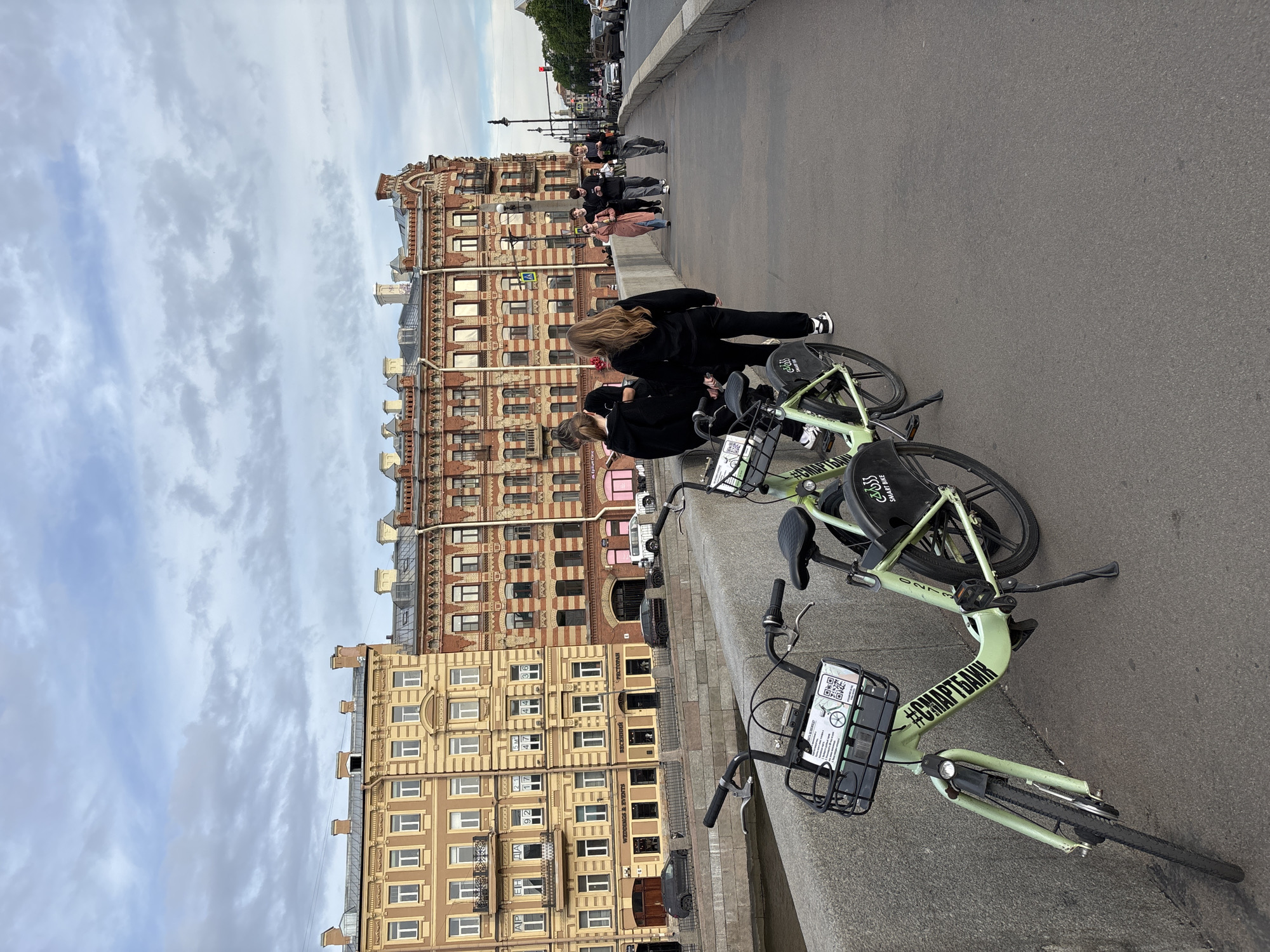This rotated portrait image captures a bustling street scene in Saint Petersburg, Russia, under an overcast sky. In the foreground, two light green rental bicycles are parked along a stone barrier. Each bike features the branding "SMART BIKE" and "#СМАРТБАНК" (Smart Bank) on its frame and a black basket equipped with a QR code and information labels. Two young women, dressed in dark clothing, stand near these bicycles, appearing to interact with their phones or the bikes. One woman has long, light-colored hair.
Beyond the bicycles, the street extends into the midground where several other people are visible, either walking or standing on the sidewalk. Some vehicles, including cars and a distinctive pink truck, can be seen in the distance, indicating an active urban environment.
The background is dominated by a row of grand, multi-story buildings with distinct European architectural styles, characteristic of Saint Petersburg. One prominent building is made of reddish-brown brick with numerous windows and decorative elements, while another adjacent building is lighter, a yellowish-beige, also featuring intricate facades and balconies. Signs with Cyrillic text, including "РЕСТОРАН" (Restaurant) and building numbers, are visible on these structures.
The sky is largely cloudy, suggesting an overcast day. The overall impression is one of everyday city life, with people utilizing a bike-sharing service amidst historical architecture.