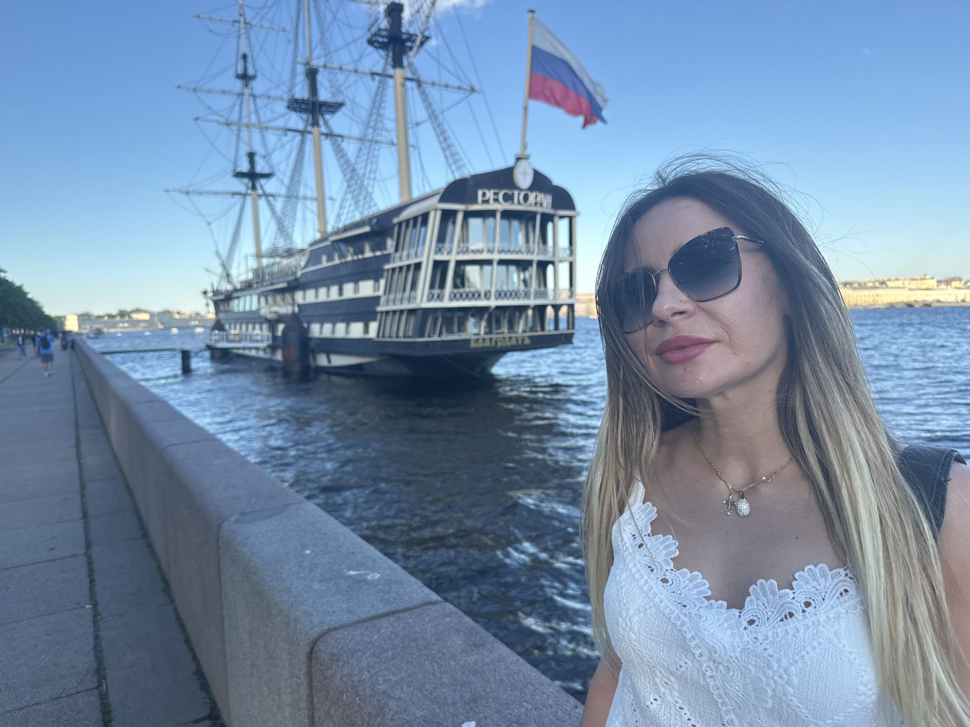 The image shows a woman standing on a quay in Saint Petersburg, Russia. Behind her, a large, dark ship with multiple masts is docked on the Neva River. The ship has a Russian flag flying from its stern and the word "РЕСТОРАН" (restaurant) visible on its upper deck. The woman is wearing sunglasses and a white lacy top, and her blonde hair is flowing. The quay is made of stone, and a sidewalk is visible to the left, with a few people walking in the distance. The background shows buildings across the river. The sky is clear and blue.