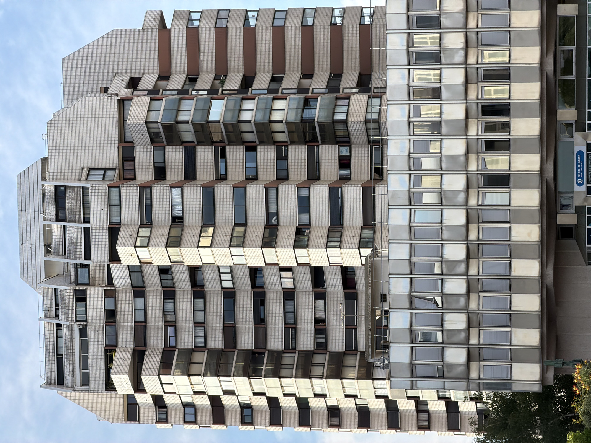 This image shows the exterior of a modern apartment building with a unique, stepped facade. The building features a textured, light-colored tile cladding and rows of windows, some of which are angled outwards. The building's design is angular and geometric, with visible balconies and exterior structural elements. On the right side of the building, there is a section with a different facade, characterized by a grid of reflective, metallic-toned windows.
A blue sign with white text is visible on the right side of the building, partially obscured. The text on the sign reads "CECCEL-VAL-DE-MARNE Chambre de commerce et d'industrie". The building appears to be located in Créteil, France, as indicated by the sign. The sky in the background is blue with scattered clouds, suggesting daytime, possibly morning or afternoon. There are no people visible in the image. The overall impression is of an urban architectural structure with a distinctive, somewhat dated, modernist aesthetic. The lighting suggests it is a clear day.