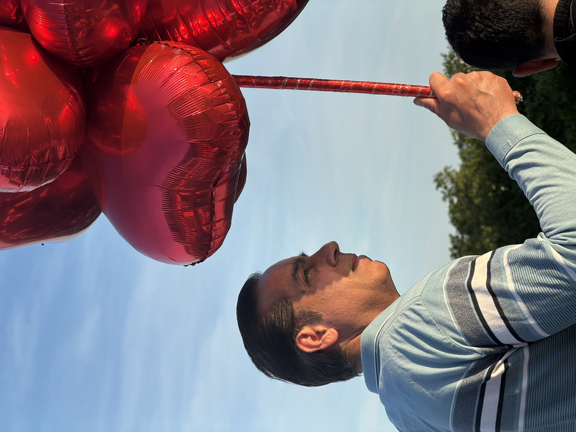 A low-angle shot shows a fair-skinned man holding a cluster of red heart-shaped balloons against a blue sky. He has dark hair, a light blue polo shirt with striped accents on the shoulders, and is looking upwards towards the balloons. His hand is gripping the red, foil-wrapped stick holding the balloons. Behind the man's hand, another person with dark hair is partially visible. The background also includes foliage, suggesting an outdoor location, possibly a park or garden. There is no information available in the picture to discern that this image was taken in Paris, France.