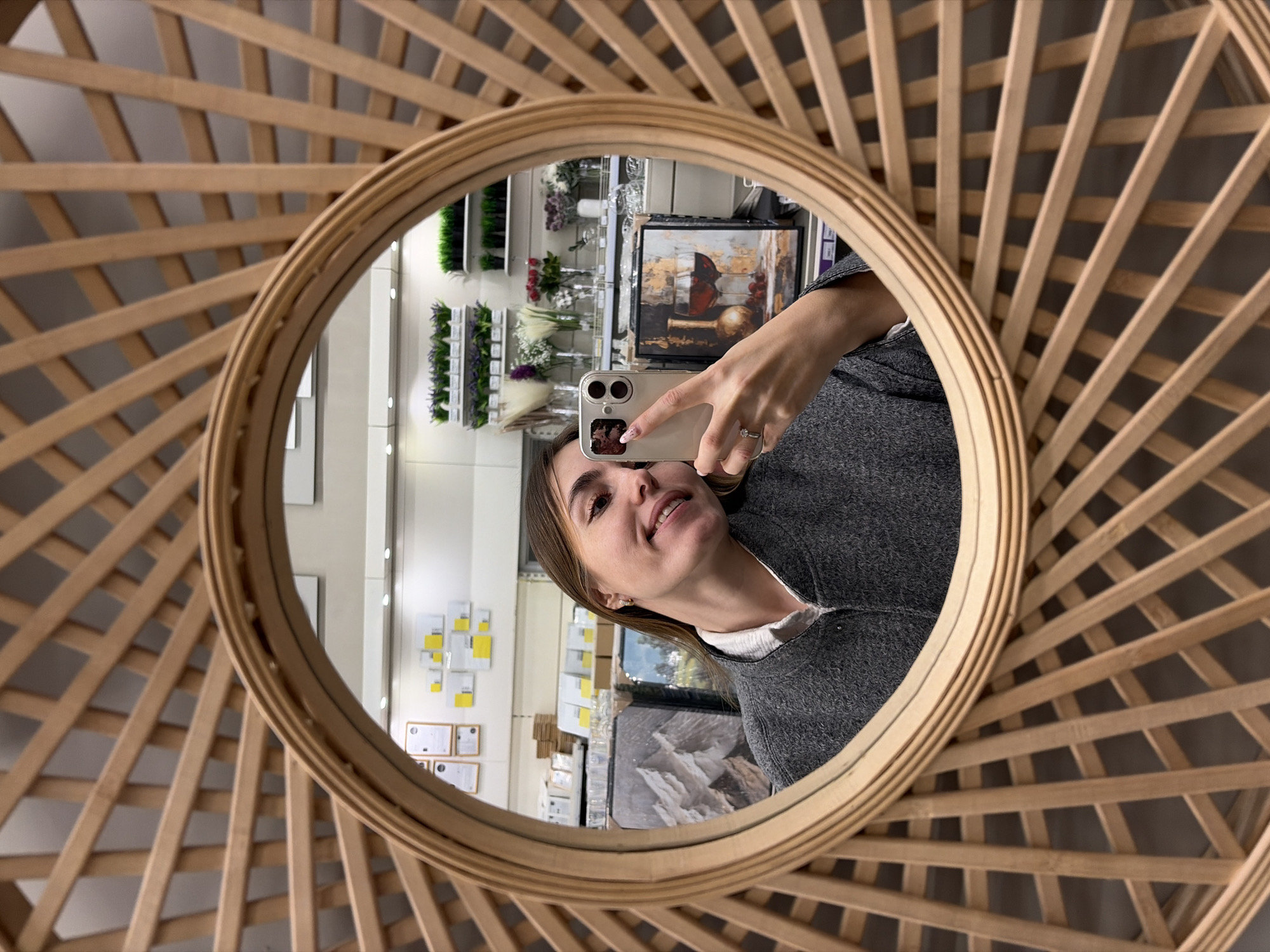 A person is taking a selfie in a round mirror with a light brown wooden frame. The frame has a sunburst design, with wooden slats radiating outwards from the mirror. The person is holding a smartphone with a tan-colored case. They are wearing a gray sweater over a white collared shirt. The mirror reflects the interior of a store. The background shows shelves with various items including plants, flowers, vases, and framed artwork. There are also papers and certificates on a wall. The location appears to be a retail establishment, possibly a home decor or gift shop. The city is not evident from the image.