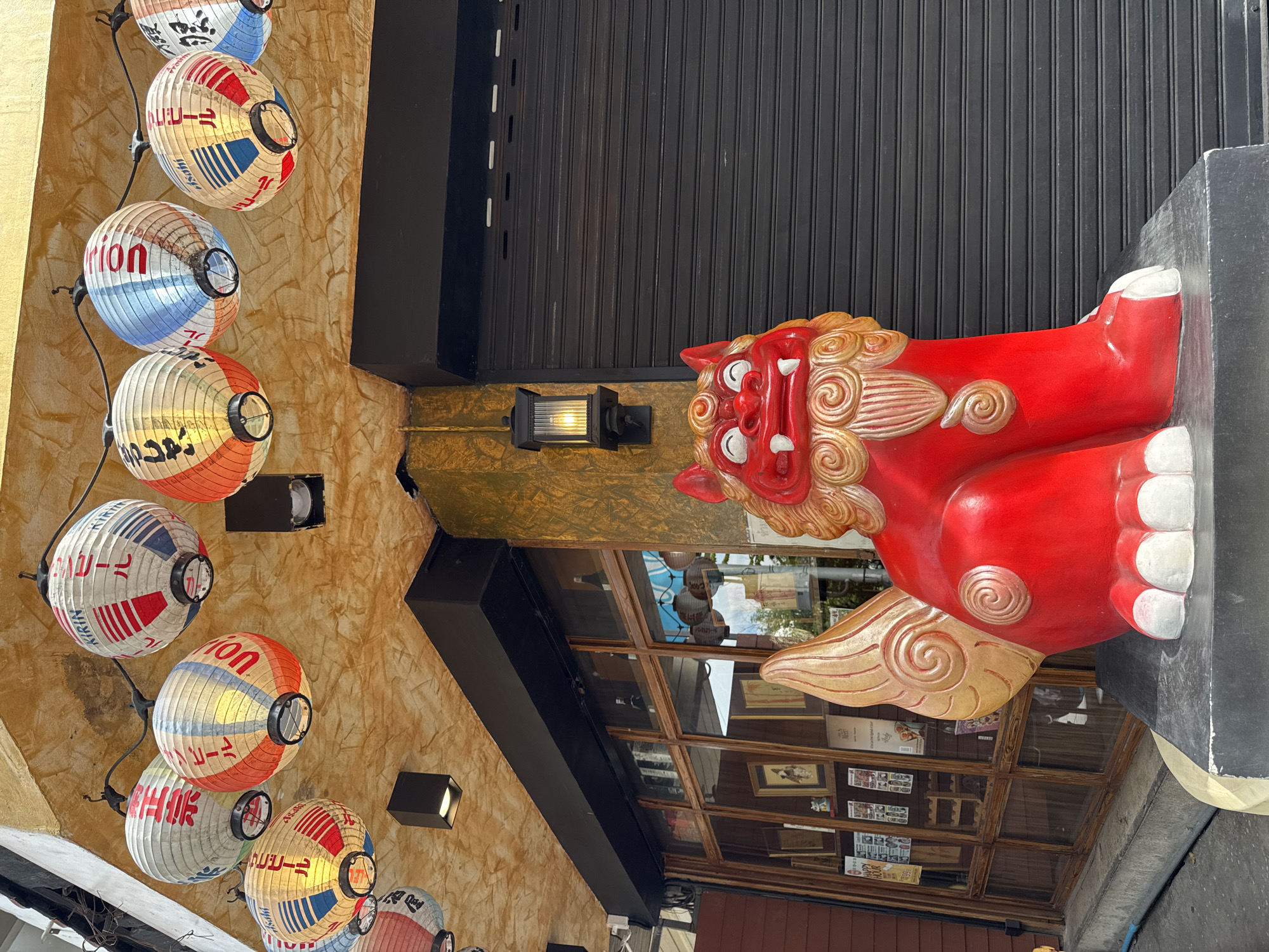 A red-and-gold Chinese lion statue sits atop a ledge outside a shop in Bangkok, Thailand. The building has a black corrugated metal awning. To the left of the lion is a series of hanging lanterns, each featuring different Japanese beer brands and colors: red, white, and blue. A modern black wall-mounted light fixture sits next to the lion's head. Through the window behind the statue, framed pictures, books, and printed documents can be seen.