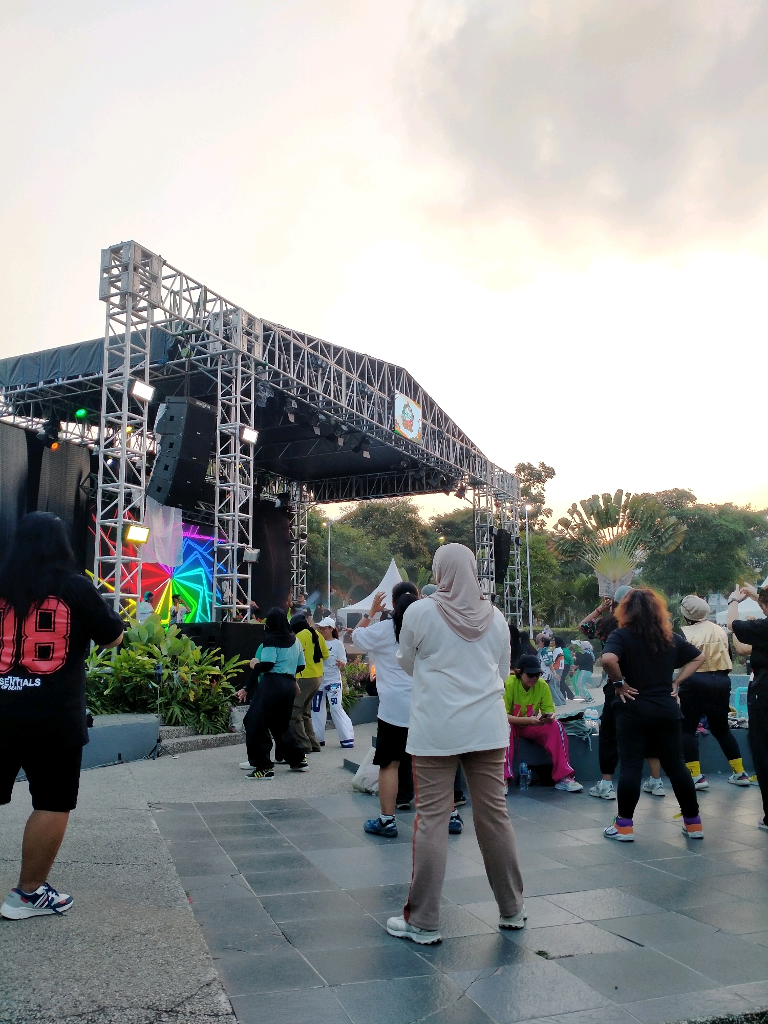 In the city of Batu, Indonesia, an outdoor stage setup is visible with a large metal truss structure supporting speakers and lights. The stage features a colorful, geometric light display. A group of people are gathered in front of the stage, some appearing to be engaged in exercise or a dance class. The foreground shows the lower bodies of several individuals, indicating they are standing on a paved area. The environment is park-like, with trees and greenery visible in the background, and the sky suggests it is late afternoon or early evening.