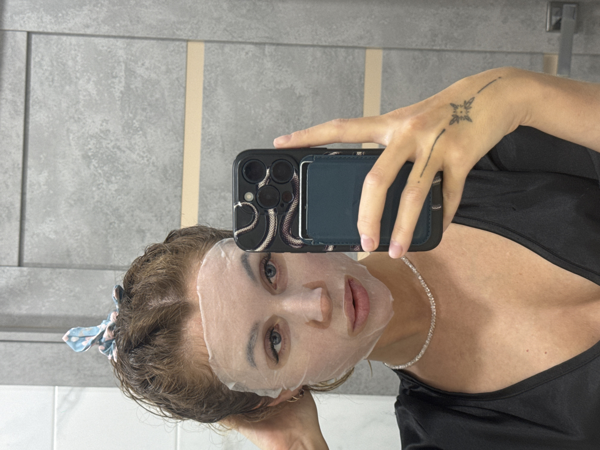 A woman is taking a selfie in what appears to be a bathroom. She is wearing a sheet face mask and holding a black smartphone with a snake-print case and a wallet attached. She has a tattoo on her hand. She is wearing a black top and a diamond necklace. A light blue hair tie is visible in her hair. The background shows a gray and white tiled wall and bathroom fixtures. The location is unknown and cannot be inferred from the image.