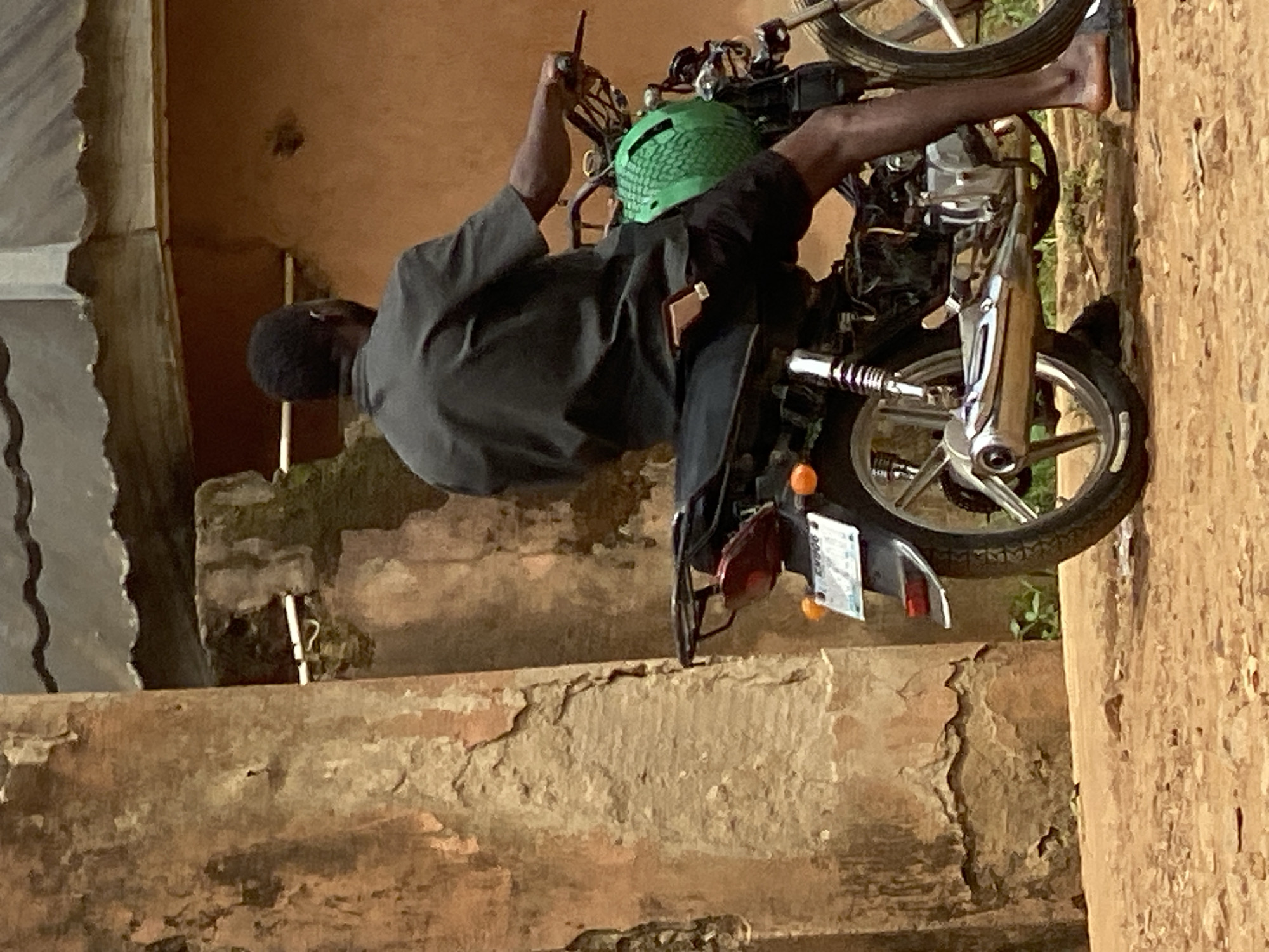 An individual is seated on a motorcycle in Maigizo, Nigeria. The individual is wearing a black shirt and a green helmet. The motorcycle is parked on a structure that appears to be either a large step or a wall, with a license plate visible at the rear. The surrounding environment shows weathered and aged walls, suggesting an older part of the city.
