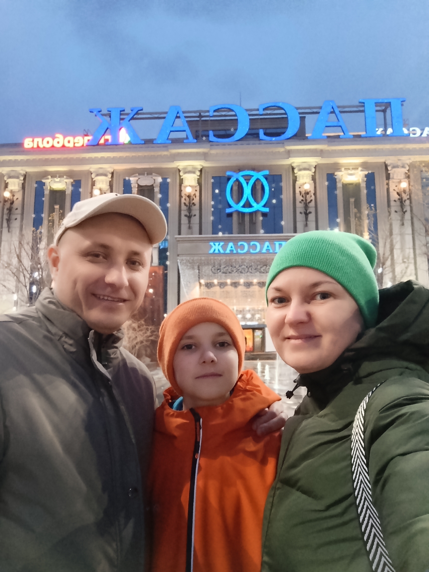 A medium shot depicts a family of three posing for a selfie. From left to right: a man wearing a grey jacket and baseball cap smiles at the camera; a child in an orange jacket and knit cap looks forward; a woman in a green jacket and knit cap smiles at the camera. All three are cropped at the mid torso. Behind them stands a building with a sign that reads "ЖАССАИ" in blue capital letters. Below the sign is an architectural detail resembling two interlocked circles. The building is further adorned with columns. The scene appears to be set in front of the business center “Continent” in Yekaterinburg, Russia. It is dusk.