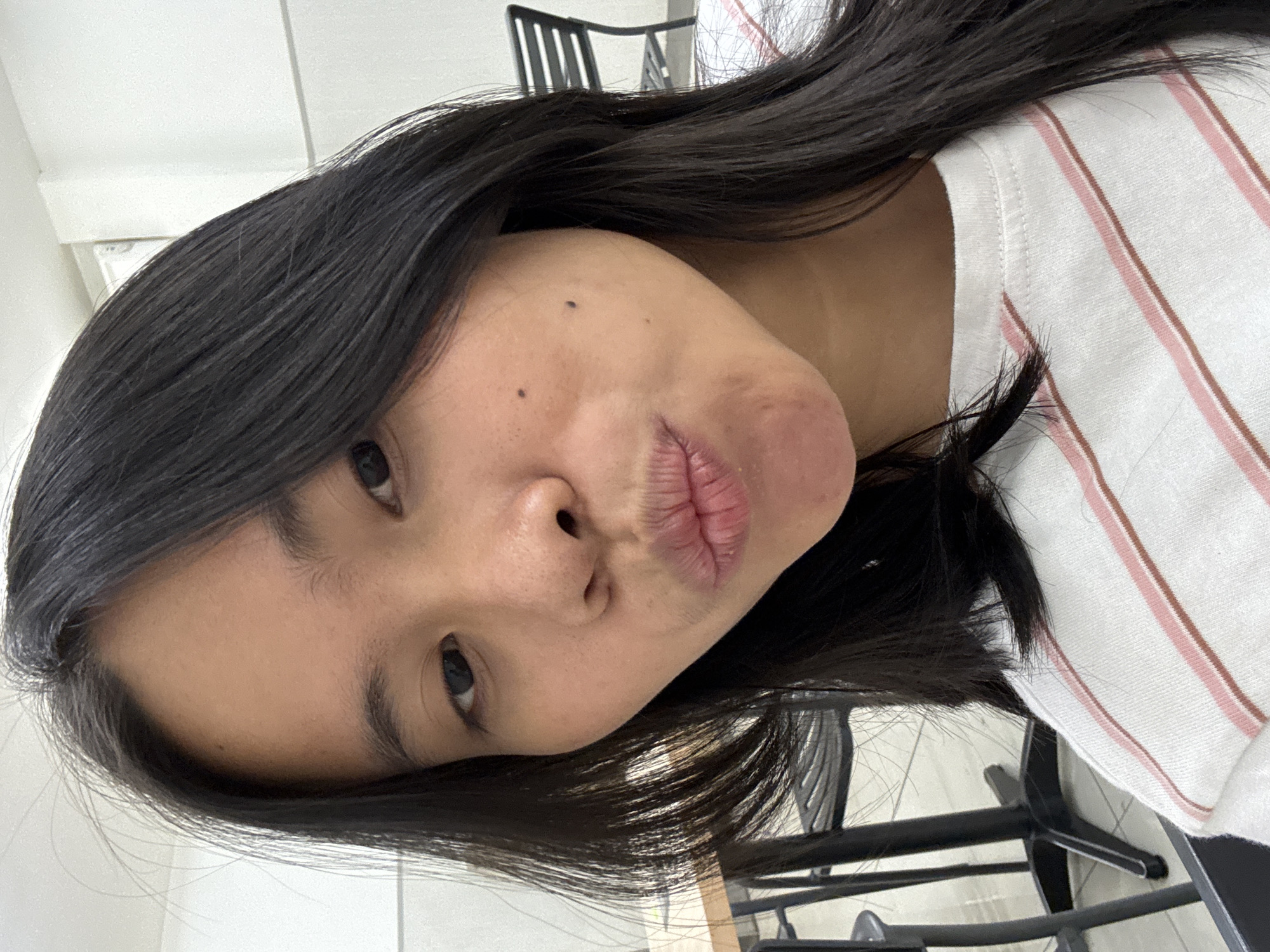 A close-up, selfie-style portrait of a young woman with long, dark hair, puckering her lips. She is wearing a white t-shirt with pink and brown stripes. The background is blurry but appears to be an indoor setting with tables and chairs, suggesting a cafe or dining area in East Jakarta, Indonesia. The lighting is bright, and the woman is the sole focus of the image.