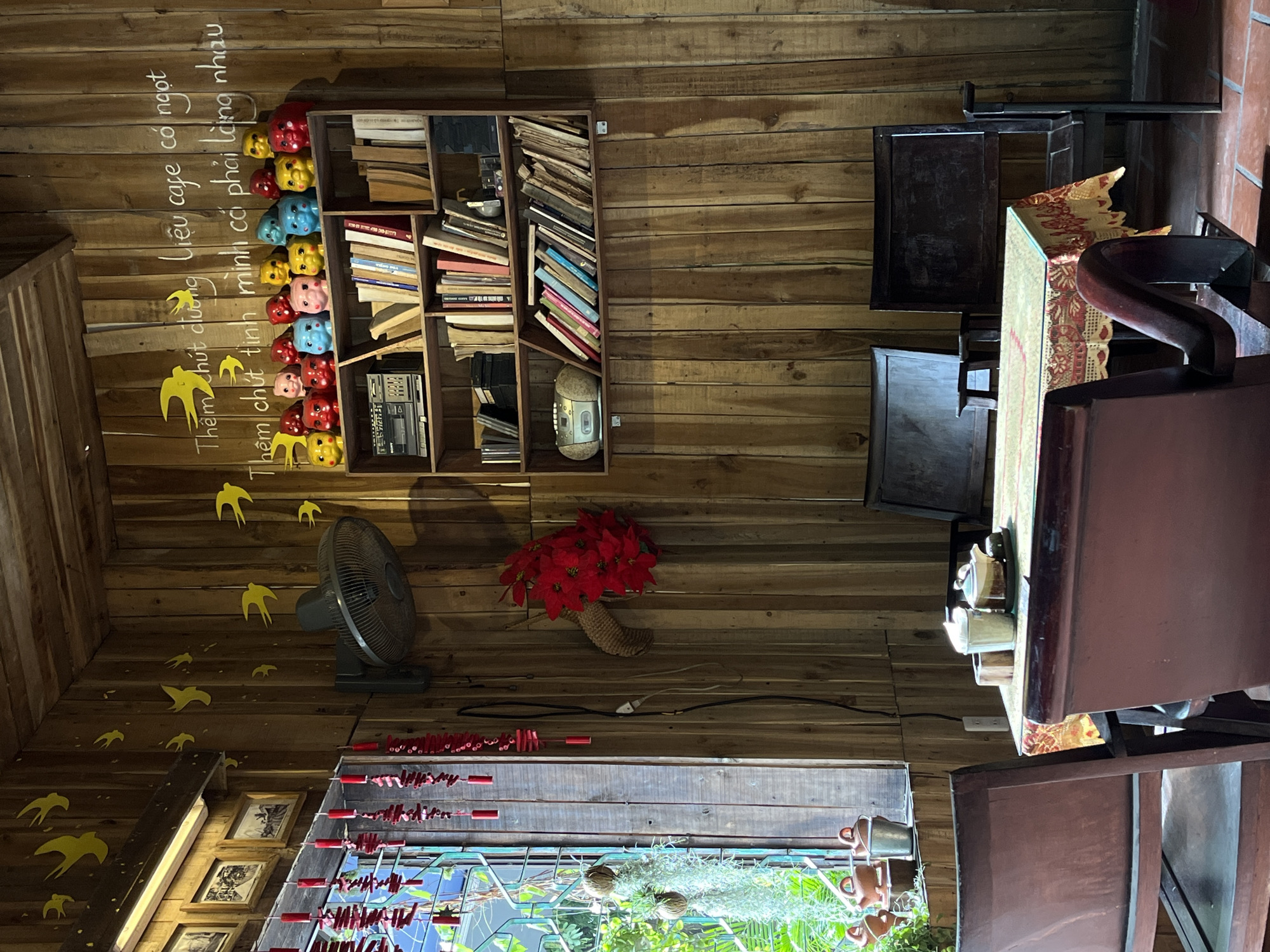 This image depicts the cozy and rustic interior of what appears to be a cafe or communal space, likely in Đà Nẵng, Vietnam. The entire room is paneled with warm-toned wooden planks, creating an inviting and traditional atmosphere. No people are visible in the scene.
On the left side, a tall wooden bookshelf is filled with numerous books, alongside a few electronic items, including a vintage-style radio or cassette player. Below the bookshelf, a playful collection of colorful decorative heads or figurines—in red, blue, yellow, and pink—are arranged. Above the shelf, white handwritten Vietnamese text is painted directly onto the wooden wall. The text reads: "Thêm chút đường liệu cafe có ngọt / Thêm chút tình mình cả phải lùng nhau," which translates to a poetic sentiment about adding sweetness to coffee and finding love. Yellow swallow-shaped cutouts are scattered across the wall, complementing the text.
In the mid-ground, a dark-colored floor fan stands ready for use. A vibrant arrangement of artificial red flowers, possibly poinsettias, rests in a woven basket. Below this, a string of red decorative items, resembling small peppers or lanterns, hangs horizontally. Framed pictures are visible on a lower wall section. Towards the bottom, a window or opening reveals green foliage and some hanging plants, suggesting an outdoor or garden view.
On the right side of the image, traditional dark wooden tables and chairs are arranged, typical of a cafe setting. One table is adorned with a patterned fabric runner featuring red and gold motifs. Another table holds a ceramic teapot and cups. The overall impression is one of warmth, cultural charm, and a peaceful ambiance, characteristic of a quaint Vietnamese establishment.
