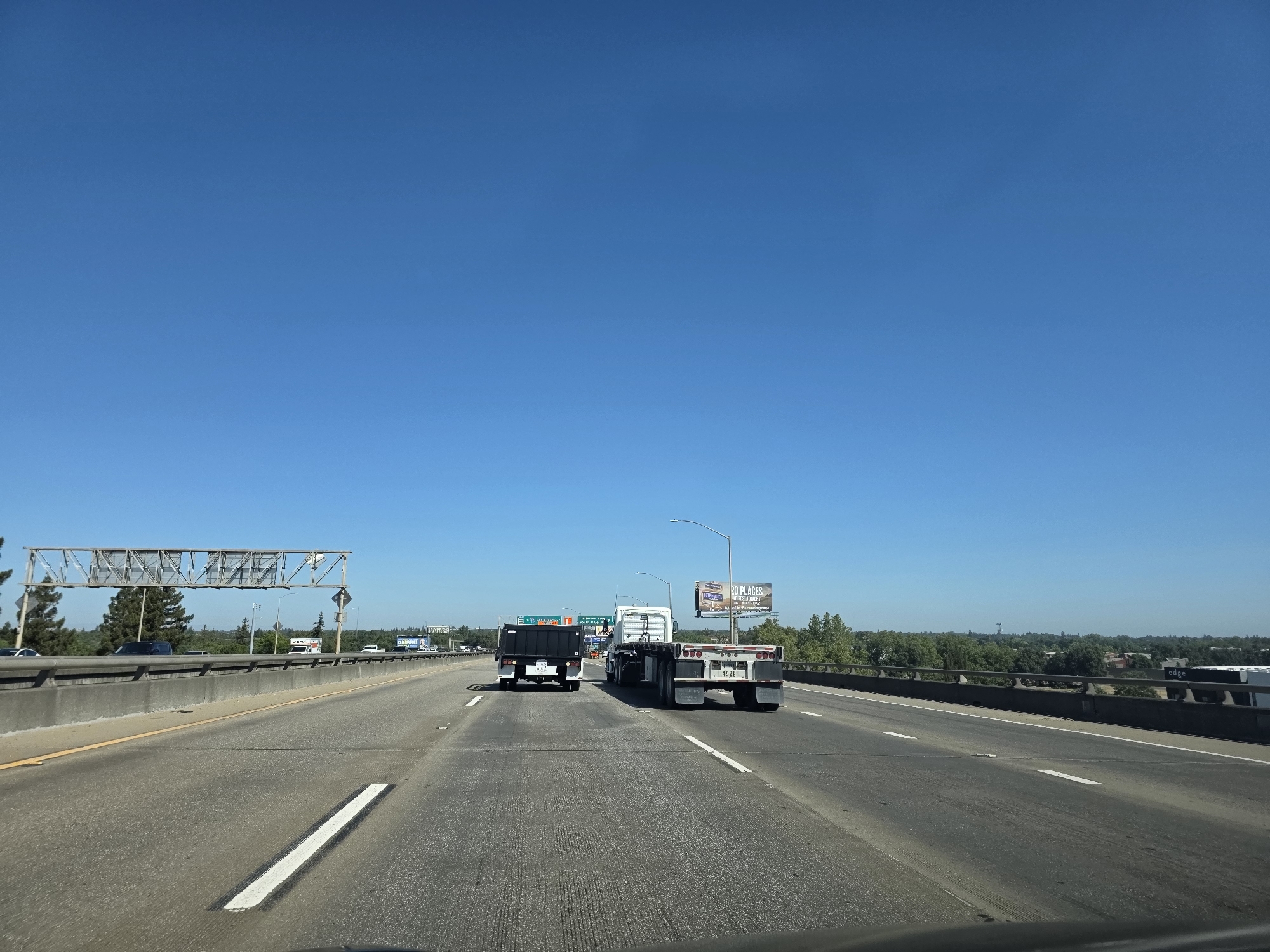 The image captures a bright, clear day on a multi-lane highway in West Sacramento, USA. The sky is a vivid, cloudless blue, indicating fair weather. The highway itself is paved and marked with white lines, with a concrete barrier on the left and a guardrail on the right separating the lanes from the surrounding environment.
Several vehicles are on the road. In the foreground, a black truck with a covered bed is in the left lane, followed by a white flatbed truck in the right lane. The flatbed truck has the license plate "4F29". Further ahead, other vehicles and trucks are visible, some disappearing into the distance. Overpasses and traffic signs are present, including a large overhead sign structure.
On the right side of the highway, beyond the guardrail, is a line of trees and some buildings. A billboard is visible in the distance, displaying the text "20 PLACES HONEST POTATOES". The overall scene suggests normal daytime traffic conditions. The perspective is from the driver's seat of a vehicle, looking forward along the highway.