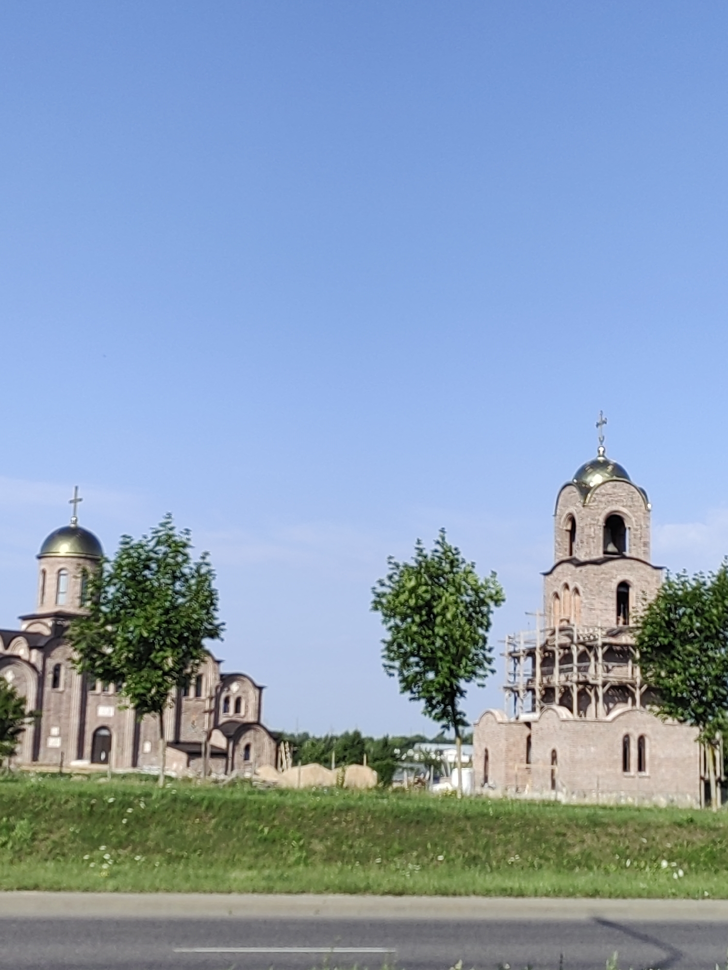 This is a photograph of two churches under construction, situated in the city of Hrodna, Belarus. The church on the left features a golden dome topped with a cross, and its brick exterior is partially visible, along with several windows and entrances. Adjacent to it is a lush green tree. The church on the right is a taller, bell tower structure, also adorned with a golden dome and cross. This structure is surrounded by scaffolding, indicating ongoing construction. The base of this church is made of brick and has several arched windows. Trees are also present in front of this building. In the foreground, a grassy embankment with small white flowers borders a paved road, which is visible at the bottom of the image. The sky is clear and blue, suggesting a sunny day.