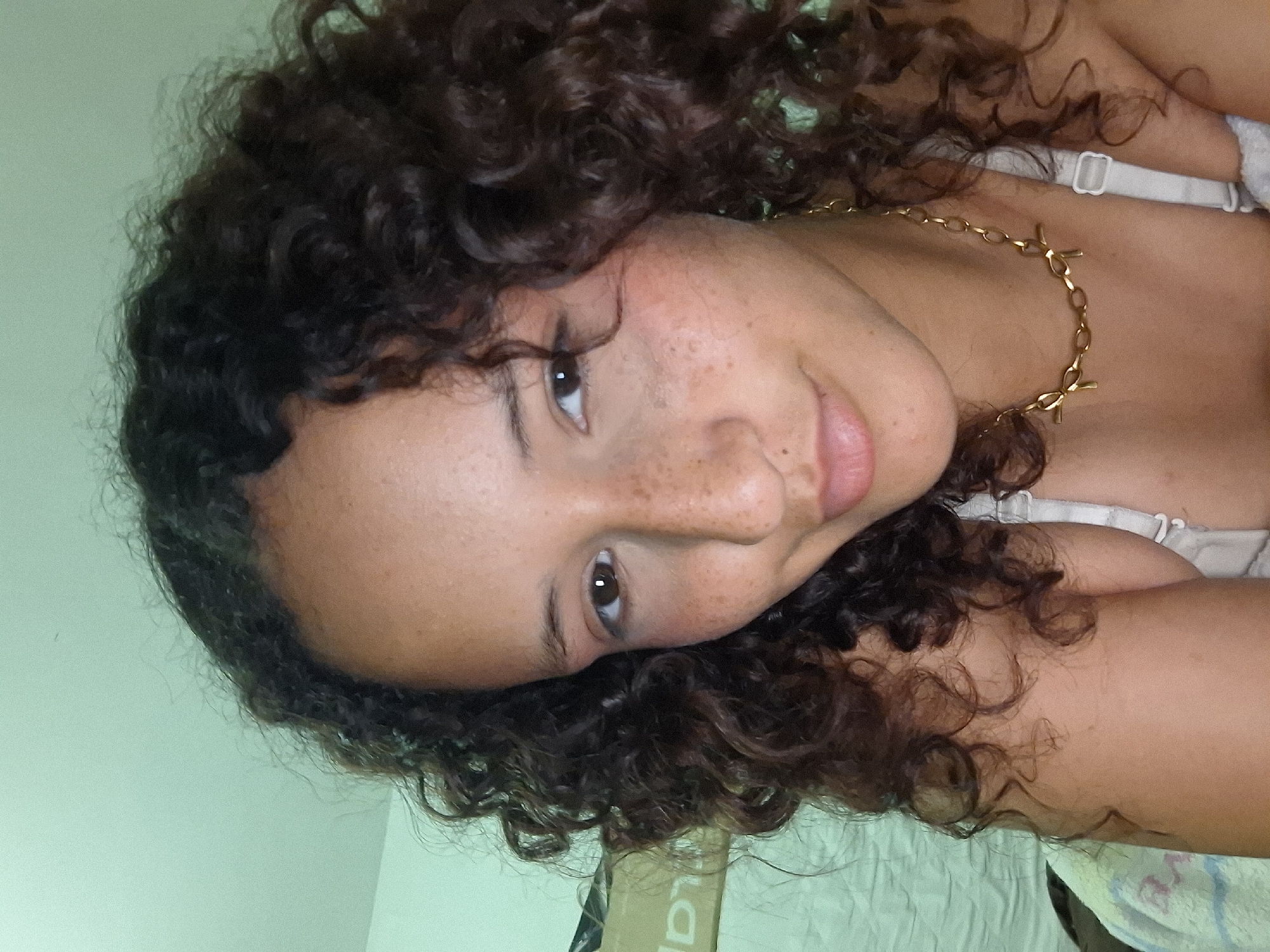 A young woman with curly dark brown hair and freckles on her face is looking at the camera. She is wearing a gold chain necklace and a light-colored bra. The background is a light green wall. The image was taken indoors, likely in a bedroom. The specific location within Juan Griego, Venezuela cannot be determined from the image.