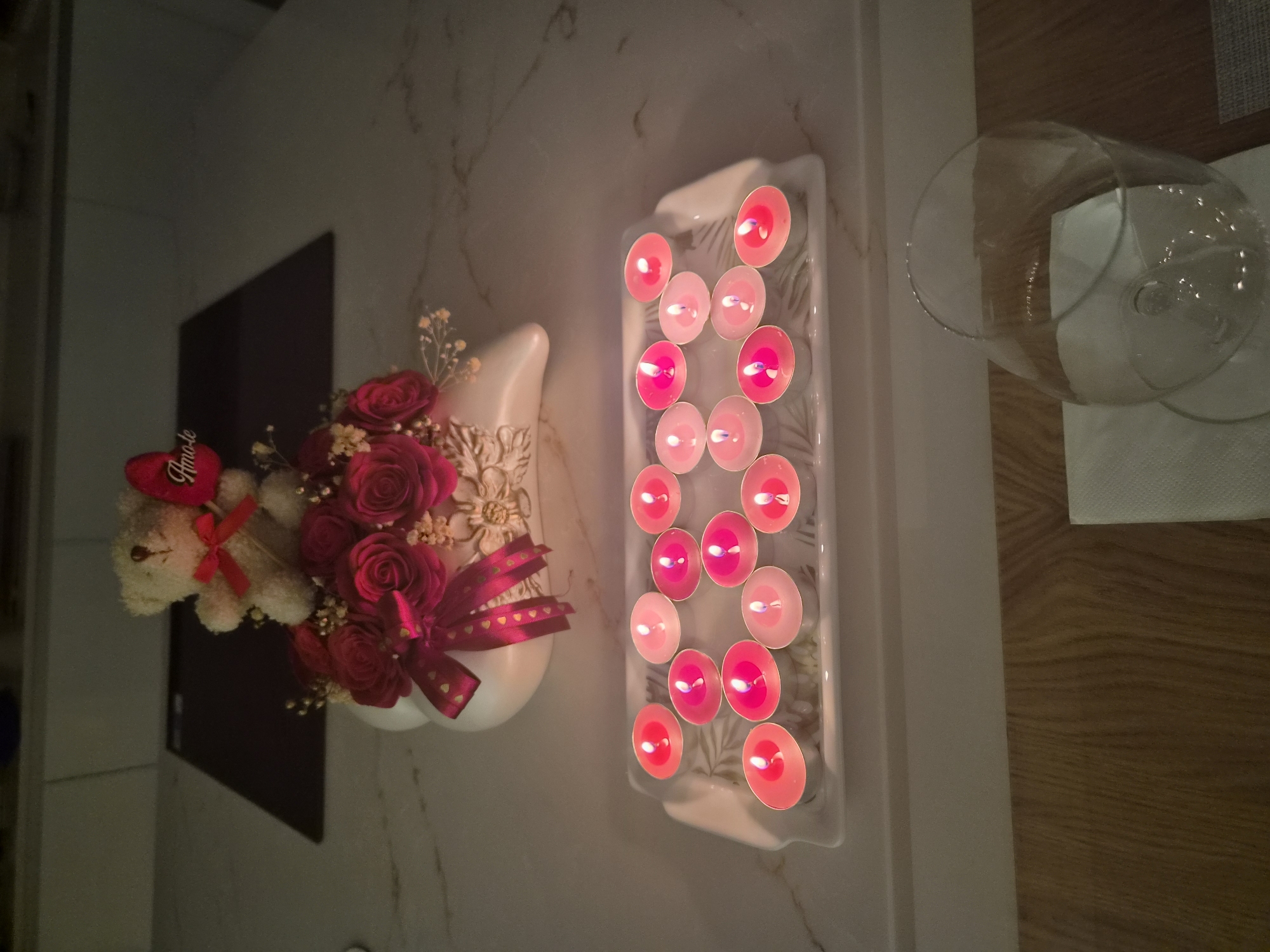 A close-up, high-angle, dimly lit shot shows a countertop with a decorative arrangement of red roses and a teddy bear. The teddy bear is white with a red bow and a red heart-shaped decoration that reads "Amo-te". The roses are arranged in a white, heart-shaped vase. In front of the vase, on a white rectangular tray with a leaf pattern, are multiple lit tea light candles in various shades of pink and red. A clear wine glass sits on a white napkin on the wooden surface below the countertop. The setting appears to be a domestic kitchen in Barcelos, Portugal.