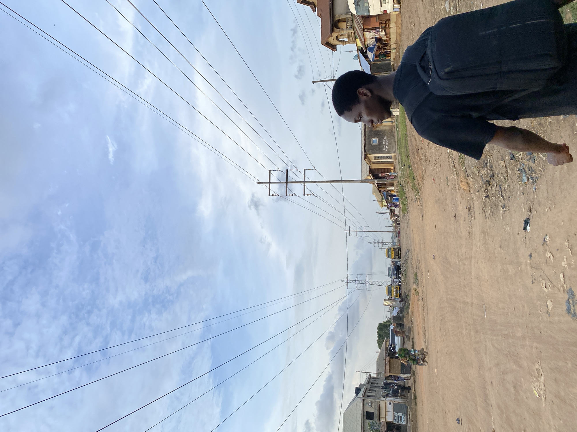 The image depicts a street in Lagos, Nigeria, characterized by a dirt road and overhead power lines extending into the distance. A dark-skinned man wearing a black shirt and backpack stands on the right side of the frame. The environment includes buildings, vehicles, and scattered debris along the roadside. The sky is cloudy.