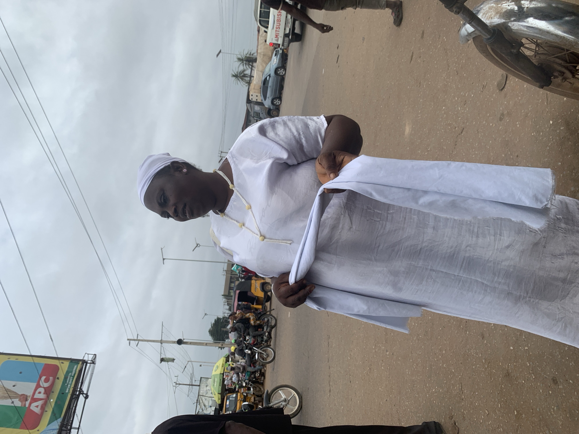 In Shagamu, Nigeria, a woman stands in a street, wearing a white headscarf and a white robe. She is holding a folded white cloth in her hands. The street is paved with dirt and gravel, and there are several motorcycles and a yellow minivan visible in the background. A billboard for the APC party is also present in the background. The sky is overcast.