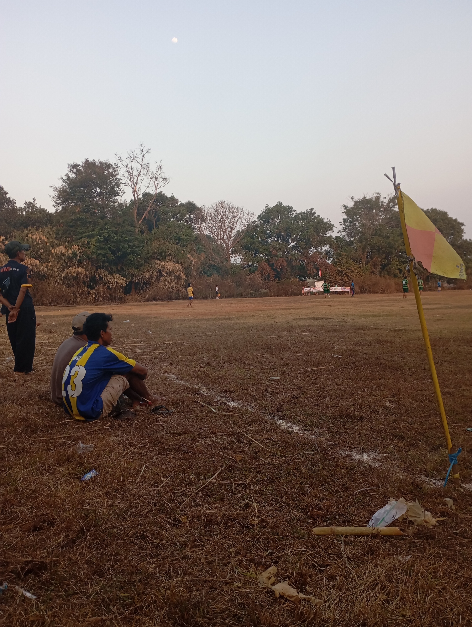 The image depicts an outdoor scene, likely a sports field, in Kecamatan Haurgeulis, Indonesia. The foreground shows dry, brown grass and scattered debris, including plastic wrappers and a piece of bamboo. Two individuals are seated in the lower left portion of the frame, facing away from the camera and towards a football match in progress on the field. One of these individuals is wearing a blue and yellow jersey with the number "3" on the back. To the left, standing and facing the field, is another person wearing a dark t-shirt and black pants, with a cap on.
The field itself is predominantly brown and appears somewhat worn, with white lines marking boundaries. In the middle ground, several figures are scattered across the field, presumably players. A goalpost with a yellow and pink flag is visible in the right foreground, attached to a yellow pole. The background consists of a dense line of trees and foliage. The sky is a pale, hazy gray, and a small, bright moon is visible in the upper left quadrant of the sky. The overall lighting suggests it is either early morning or late afternoon.