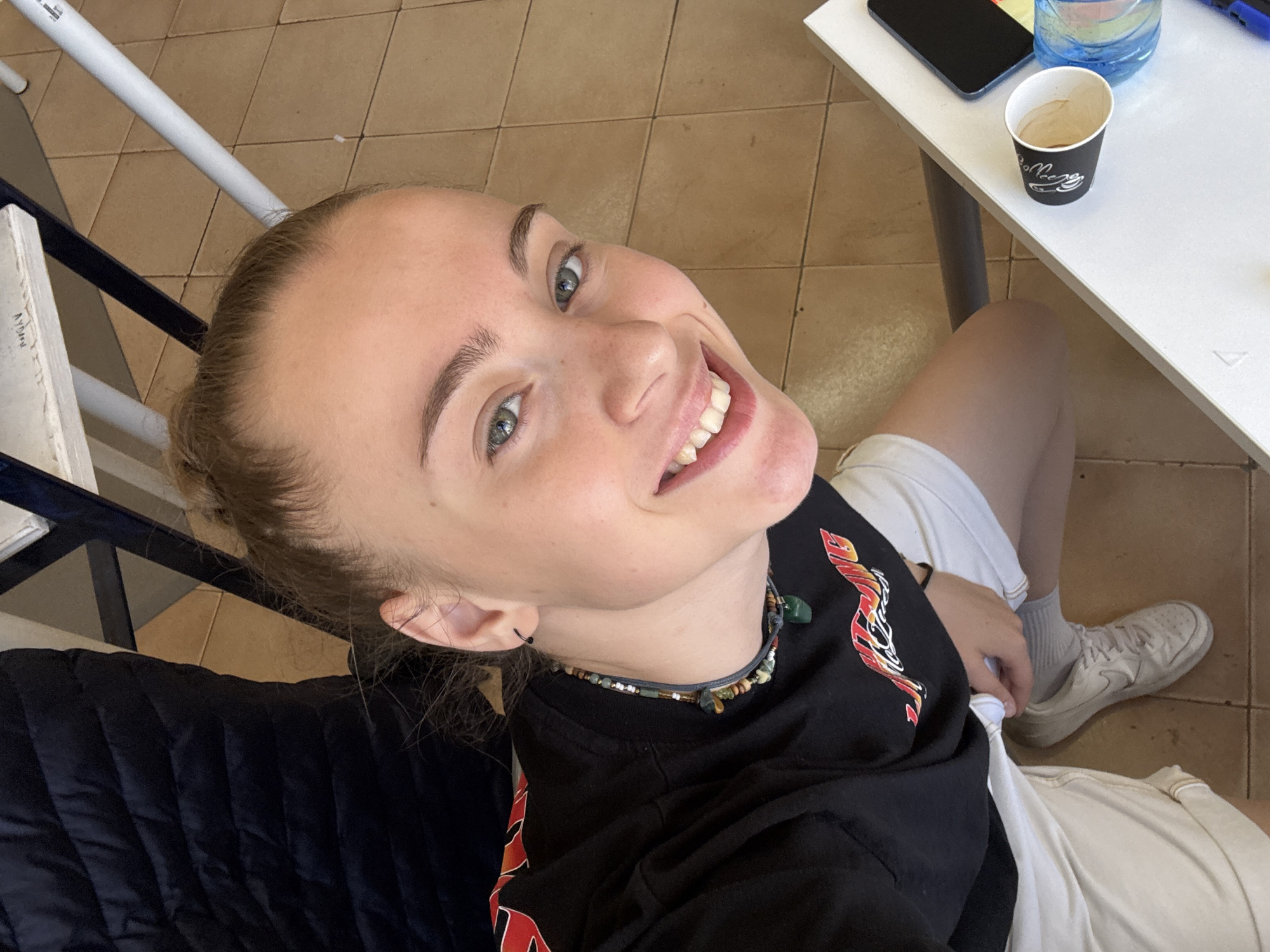 A young woman with blonde hair pulled back in a bun smiles at the camera. She is wearing a black t-shirt with a red and yellow design on the sleeve, and white shorts. She has a necklace made of beads and stones. Her left ear is visible, with a small black hoop earring. She is sitting at a white table. On the table, there is a black smartphone, a white paper cup with a black logo, and a blue plastic water bottle with colorful liquid. Her feet are visible, wearing white sneakers and grey socks. The background shows a tiled floor and what appears to be furniture. The scene is well-lit, suggesting it is daytime. The overall impression is casual and relaxed.
