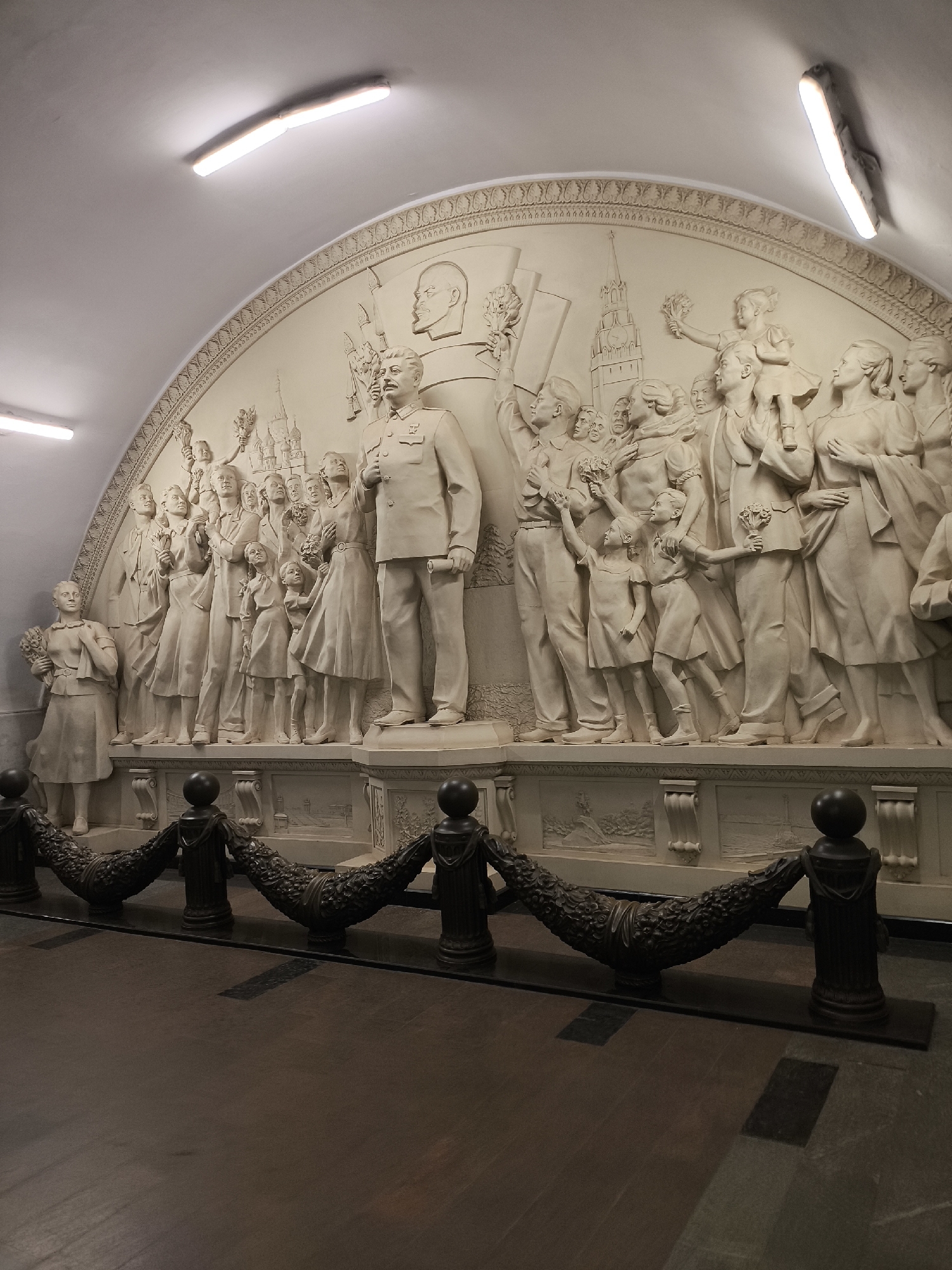 The image shows a large, ornate bas-relief sculpture depicting a procession of people, including children, holding flowers and looking upwards. The sculpture is made of light-colored stone and is set within an arched wall, illuminated by fluorescent ceiling lights. A prominent male figure in a military uniform stands at the right of the sculpture. The sculpture appears to be located in a public space, possibly a metro station, given the presence of a dark wooden floor in the foreground and a decorative metal barrier with dark posts and ornate rope-like elements. Architectural details, including decorative moldings and smaller relief panels, are visible along the base of the sculpture and the wall. The overall scene conveys a sense of celebration and collective movement.