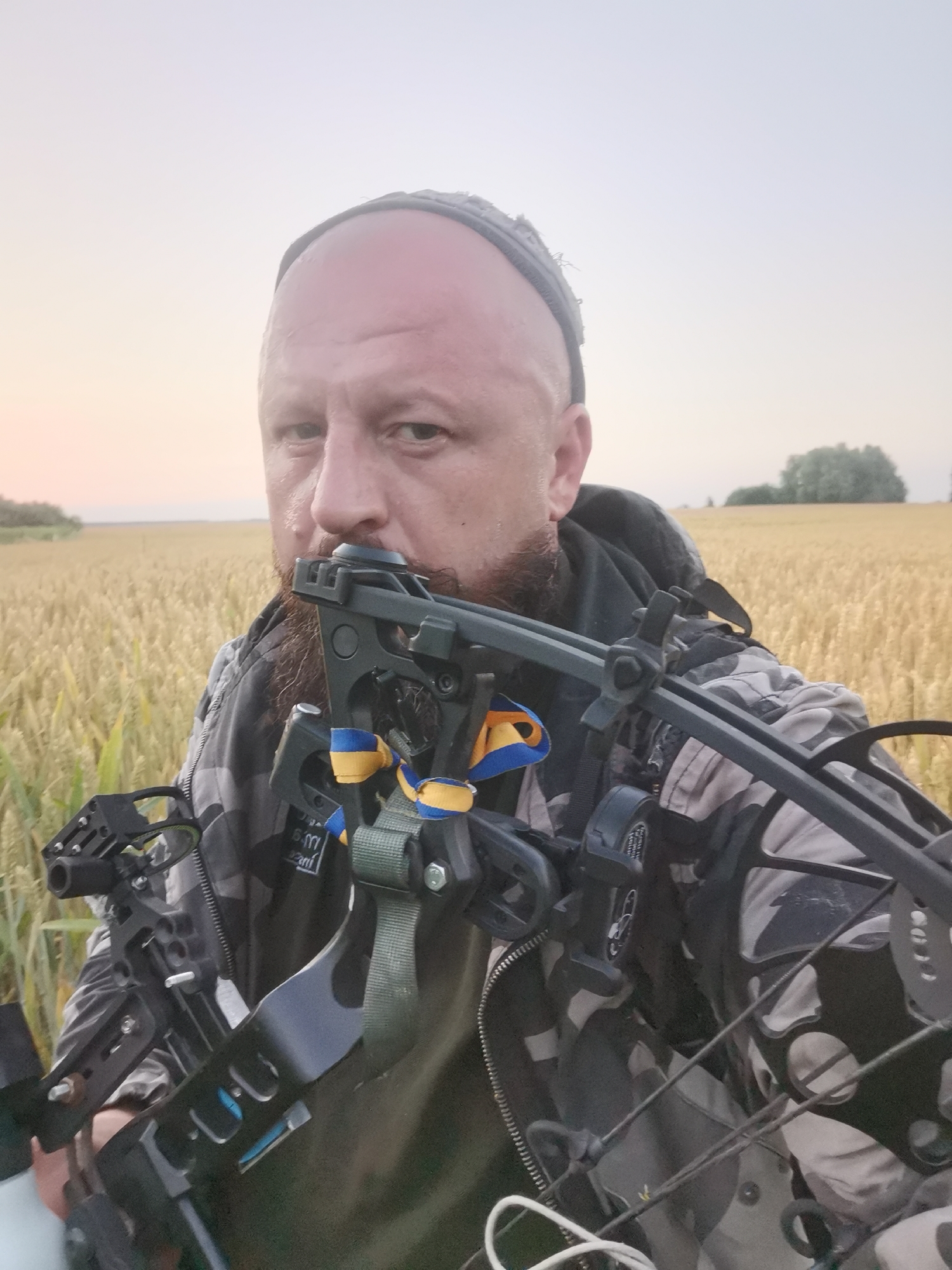 A man with a shaved head and a beard is taking a selfie in a field of wheat. He is wearing a camouflage jacket and a dark beanie. He is holding a compound bow across his chest. The bow has a stabilizer with yellow and blue ribbons attached, which are the colors of the Ukrainian flag. The background is a blurred field of golden wheat under a pale sky. The location is identified as Semypolky, Ukraine.