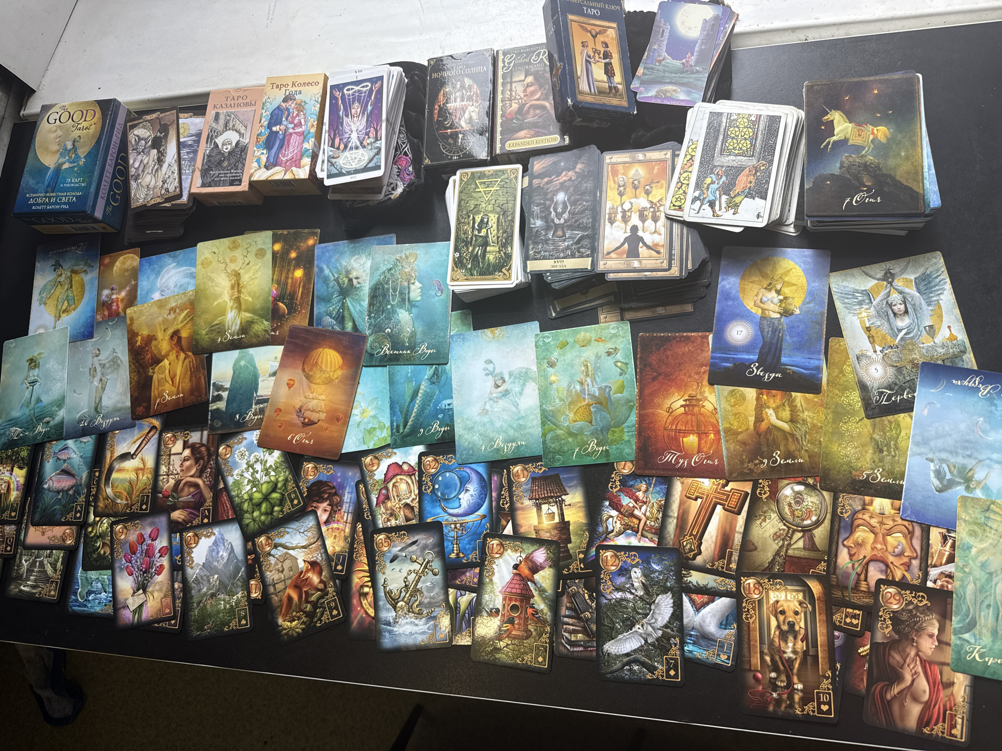 The image shows a collection of tarot and oracle card decks and individual cards spread out on a black surface. Some decks are still in their boxes, including "The Good Tarot" by Colette Baron-Reid, "Таро Казановы" (Tarot Casanova), and "Таро Колесо Года" (Tarot Wheel of the Year). Several other decks are visible but not clearly identifiable. A large number of cards from different decks are spread out, face up, revealing their artwork. The cards display various images, including people, animals, landscapes, and symbolic elements. There is no clear indication that the location is in Saint Petersburg, Russia, and there are no people present in the image.