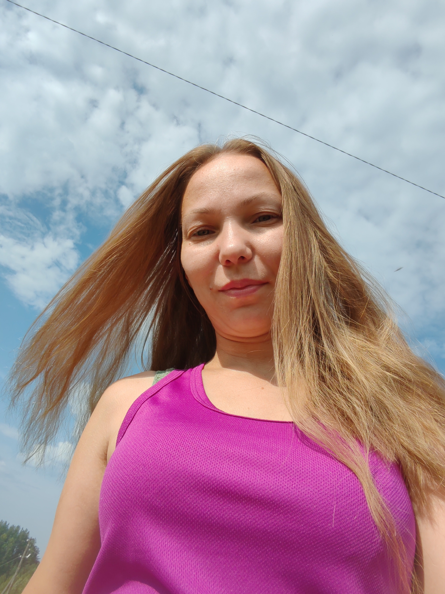 A woman with long blonde hair is pictured from a low angle, looking up towards the camera. She is wearing a purple tank top. The background is a bright blue sky with scattered white clouds. A thin dark wire stretches diagonally across the upper portion of the image. There is a hint of green foliage and trees at the very bottom of the frame, suggesting an outdoor location. The image appears to be a selfie taken during daylight.