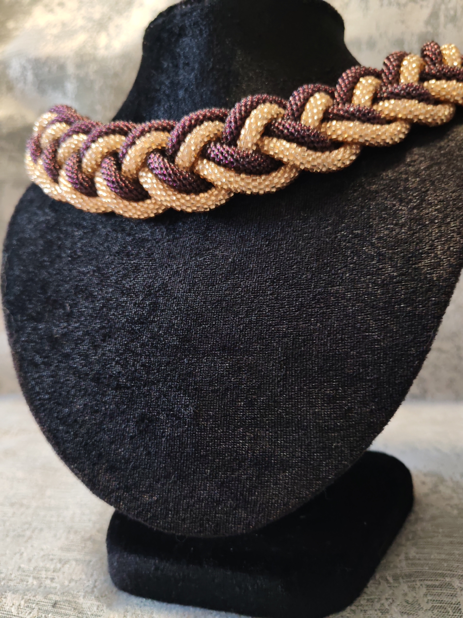 A beaded necklace with gold and purple strands is displayed on a black velvet bust. The necklace features a braided pattern. The bust is on a textured surface, likely a fabric. The background is a blurred, textured gray fabric. The scene is well-lit, with soft shadows. The location is unknown.