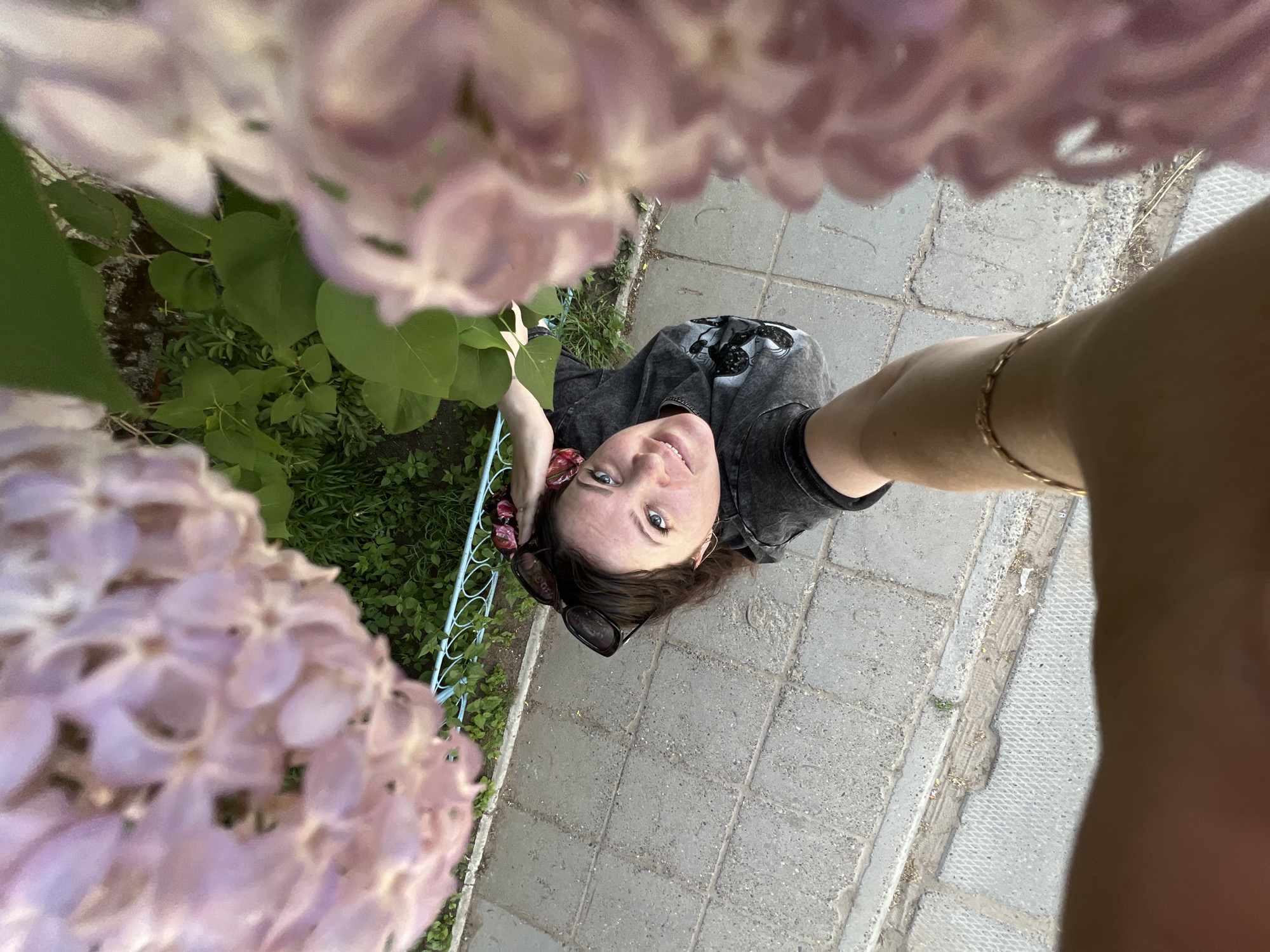 A woman is looking up at the camera, which is positioned at a low angle, while standing on a paved area. The woman has fair skin, dark hair, and is wearing a dark t-shirt. Sunglasses are perched on her head, and she is adorned with a bracelet on her arm. Lilac bushes with green leaves surround her. The paved area is composed of gray bricks. The location is implied to be in Noviy, Russia.