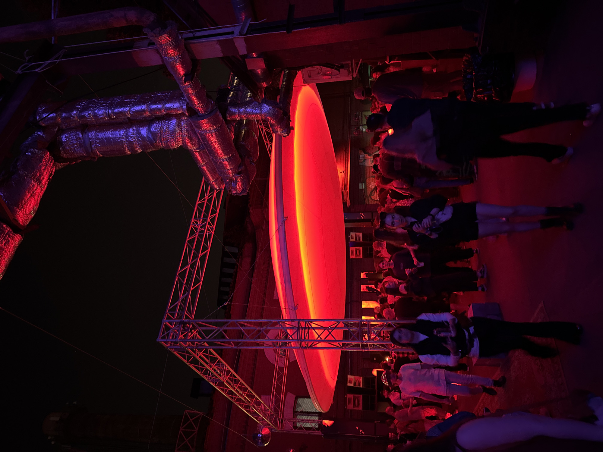 This is a nighttime photograph depicting a large gathering or event held within an industrial-style venue in Moscow, Russia. The scene is dominated by a prominent, circular, luminous red fixture suspended from a metal truss system, casting a strong crimson and deep pink glow across the environment. Above and to the right, an extensive network of insulated metal ductwork and piping is visible, also illuminated with red light, creating reflective surfaces. A smaller metal truss structure extends towards the upper left, from which a small disco ball hangs. In the dark background, silhouettes of urban buildings are discernible, including one with a tall, cylindrical or turret-like feature.
Approximately 50 to 70 individuals, primarily adults, are present across the foreground and mid-ground. They are casually dressed in various attire, including t-shirts, vests, and pants, and are engaged in activities such as walking, standing, and conversing. Many faces are partially obscured by shadows and the intense red lighting. Some individuals are holding beverages or mobile phones. Black rectangular speakers are mounted on vertical supports near the crowd. The ground appears to be a dark, paved surface, with a dark red patterned rug visible in the mid-ground. Various electrical conduits and boxes are affixed to vertical structural elements on the right side.