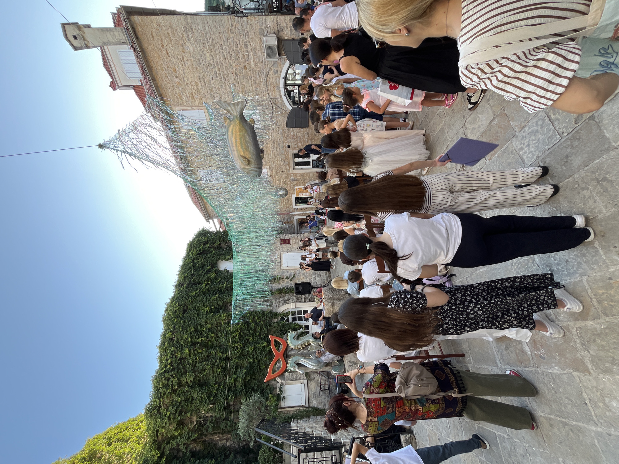 Here's a detailed description of the image:
The image captures an outdoor event in what appears to be a public square in Budva, Montenegro. The scene is oriented vertically.
**Environment:**
The event is taking place in a stone-paved square. Buildings constructed of stone are visible, one covered in greenery. Above the event, a decorative net hangs with a large sculpture of a fish. A large orange mask sculpture can be seen as well.
**People:**
A large crowd is present, with many people seated and facing a central point, suggesting an event or performance. Many are wearing white shirts. Several children are present. The crowd is observing the presentation.
**Objects:**
A stage is set up at the center. Speakers can be seen on the right of the image.
**Overall Impression:**
The image gives the impression of a public celebration or performance taking place in a historic urban setting. The presence of children and the decorations indicate a community-oriented event.