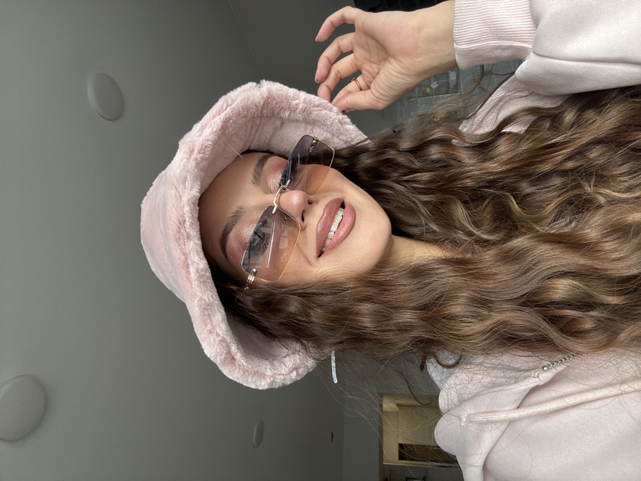 A female individual is depicted in a medium close-up, indoors. She has long, wavy brown hair extending over her shoulders and chest. She is wearing a pink faux fur bucket hat and rimless rectangular sunglasses with gradient amber-to-darker lenses and gold-colored bridge and temple accents. Her lips are slightly parted in a smile, revealing teeth, and coated with a light pink gloss. Reddish-pink eyeshadow is visible on her eyelids. She wears a light pink hooded sweatshirt with a silver-toned zipper. Her left hand is raised near the left side of her head, fingers slightly curled, appearing to touch or adjust her hat or hair. The background is a blurred indoor setting, consisting of a light-colored wall and ceiling. A round, white ceiling light fixture is visible in the upper background. A blurred doorway or opening with a visible frame is discernible on the far left. The specified location of Krasnoyarsk, Russia, cannot be independently verified from the visual information provided within the image.