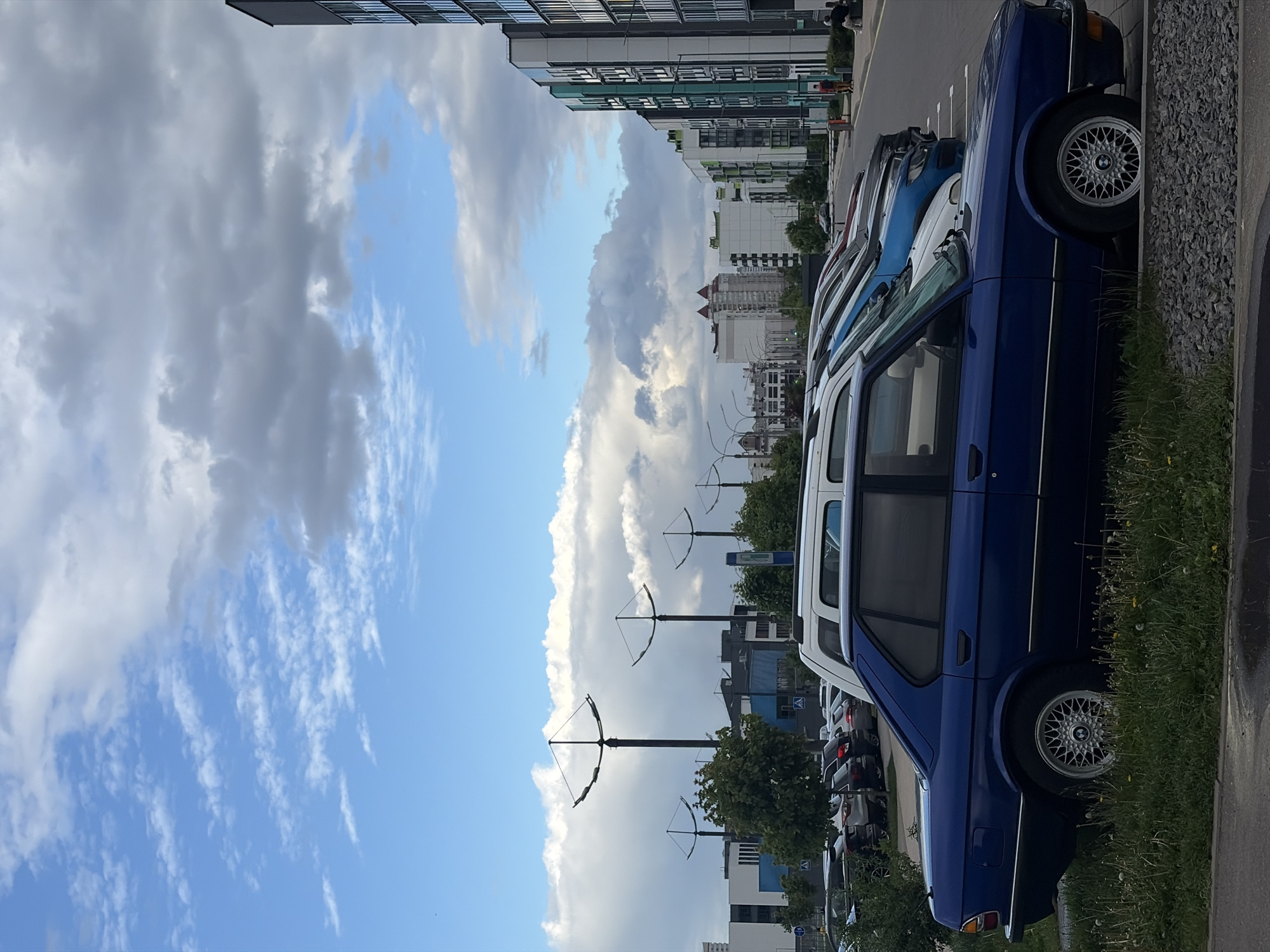 The image shows a row of cars parked on a street in Бараўлянскі сельскі Савет, Belarus. The foreground contains a blue and white vintage BMW 5 series. The tires have a silver, lattice-like pattern. Behind the BMW are several parked cars of various colors, and in the background is a modern building with green and white cladding. Further in the background is a residential area and a partly cloudy sky. Black lamp posts with decorative bow-shaped lighting fixtures are visible in the middle ground. The photo is taken from a slightly low angle.