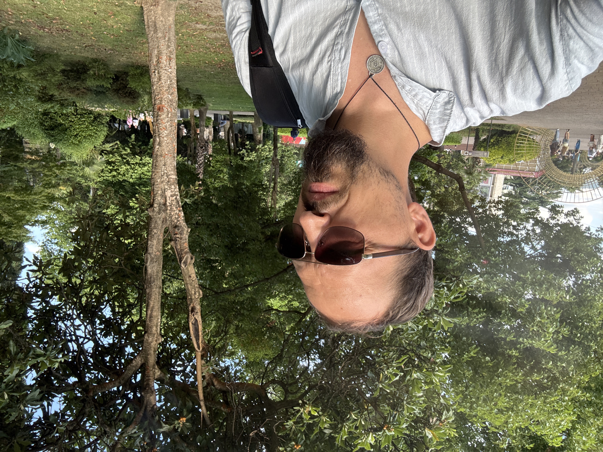 The image is a close-up selfie of a man with a beard and sunglasses, taken outdoors. He is wearing a light-colored collared shirt and a black backpack. A necklace with a circular pendant is visible around his neck. The background features lush green trees and foliage, suggesting a park or natural setting. In the distance, there are some structures that appear to be part of an outdoor exhibition or attraction, possibly a large, abstract sculpture made of golden material. A few people are visible in the background, appearing to be walking around. The lighting suggests it is daytime, possibly with overcast skies or shade from the trees. The overall mood appears casual and relaxed. The image is oriented sideways.