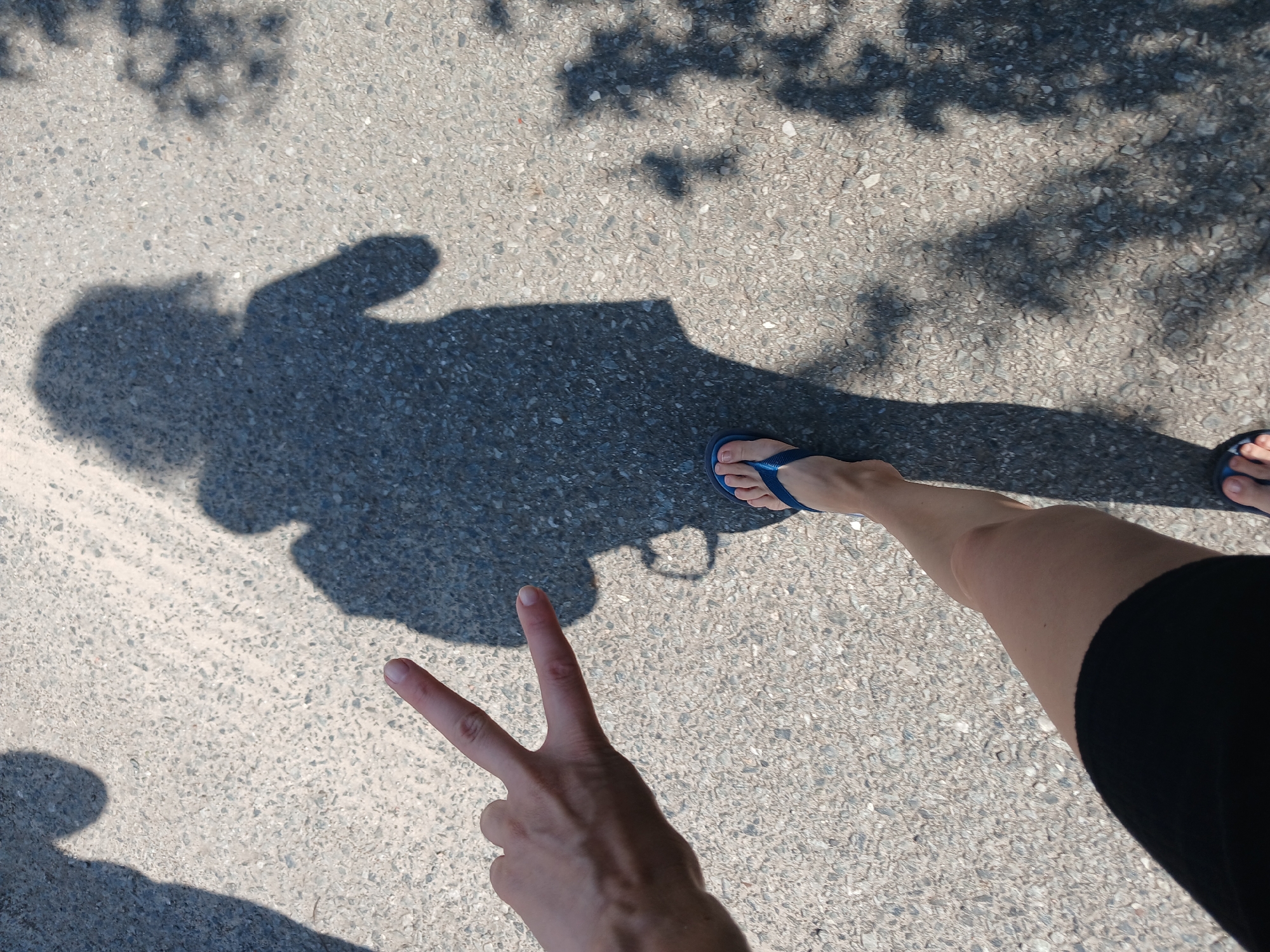 In an overhead shot, a person's legs and arm are visible on a textured asphalt surface, casting a shadow. The person is wearing black shorts and blue flip-flops. Their left arm is extended, with fingers forming a peace sign. The shadow suggests the person is carrying a backpack. Other shadows, possibly from trees and another person, are also present on the ground. The scene appears to be outdoors, likely on a paved pathway in the city of Maslova Pristan', Russia.