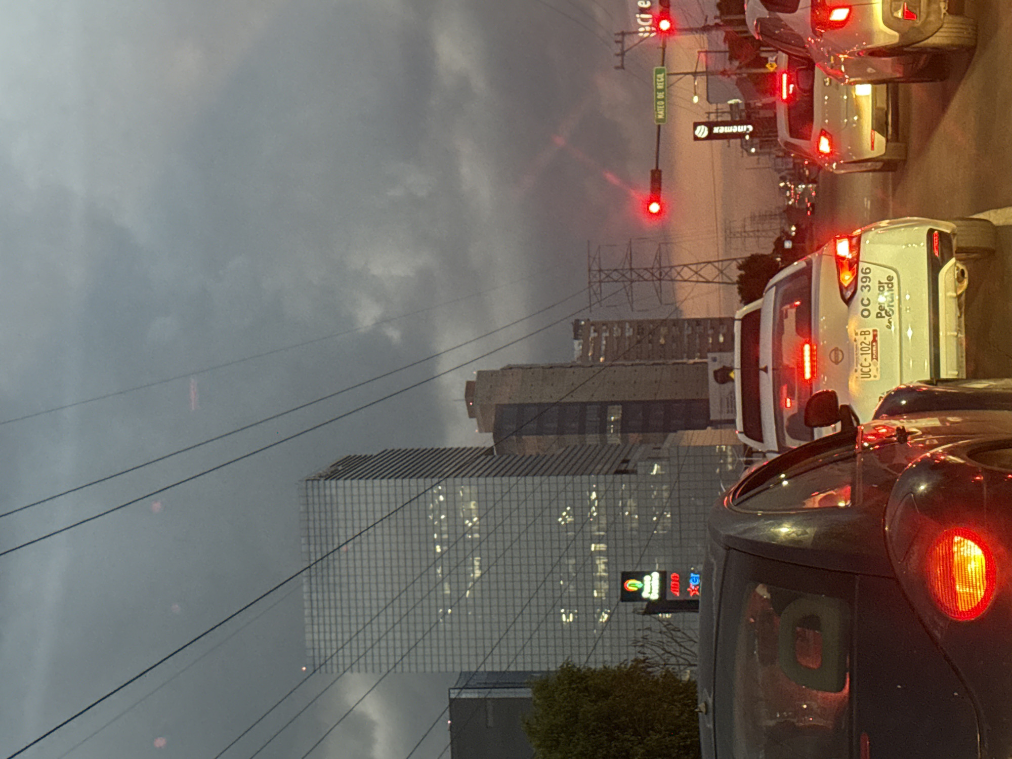The image shows a view from inside a vehicle, likely at a standstill in traffic, looking towards the city of Puebla, Mexico. The scene takes place at dusk under an overcast sky. The traffic signal displays a red light. A tall, modern building with a grid-like facade dominates the view. A smaller building behind the grid building features signage including "OXXO Gas." A sign for "MATEO DE REGIL" street can be seen attached to the traffic light pole. Several vehicles, with their brake lights illuminated, are stopped ahead. A white vehicle displays the license plate "UCC-102-B OC 396". A Cinépolis sign is present as well.