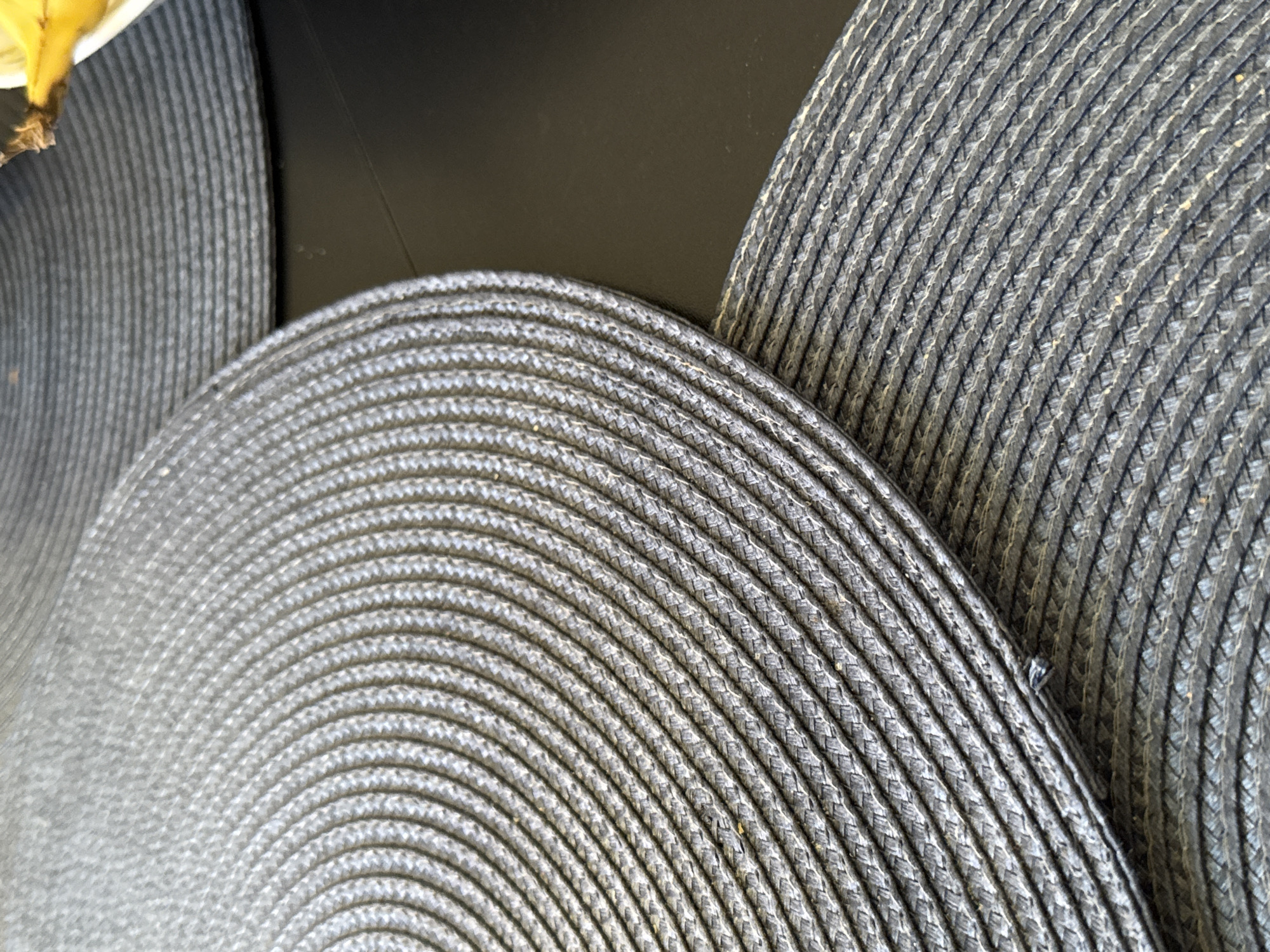 This is a close-up, overhead shot of two round, woven placemats on a dark surface. The placemats are made of a material that appears to be some kind of synthetic fiber, woven in concentric circles. The texture is rough and detailed, showing individual threads. The placemats are dark gray in color and are overlapping each other, with the larger one in the foreground partially obscuring the smaller one behind it. The background surface is a solid dark color, likely a table. A small portion of what appears to be a yellow banana and a white bowl is visible in the upper right corner, suggesting this might be a dining setting. The lighting is natural and comes from above, casting subtle shadows that highlight the texture of the placemats. The image captures a domestic scene, likely indoors.