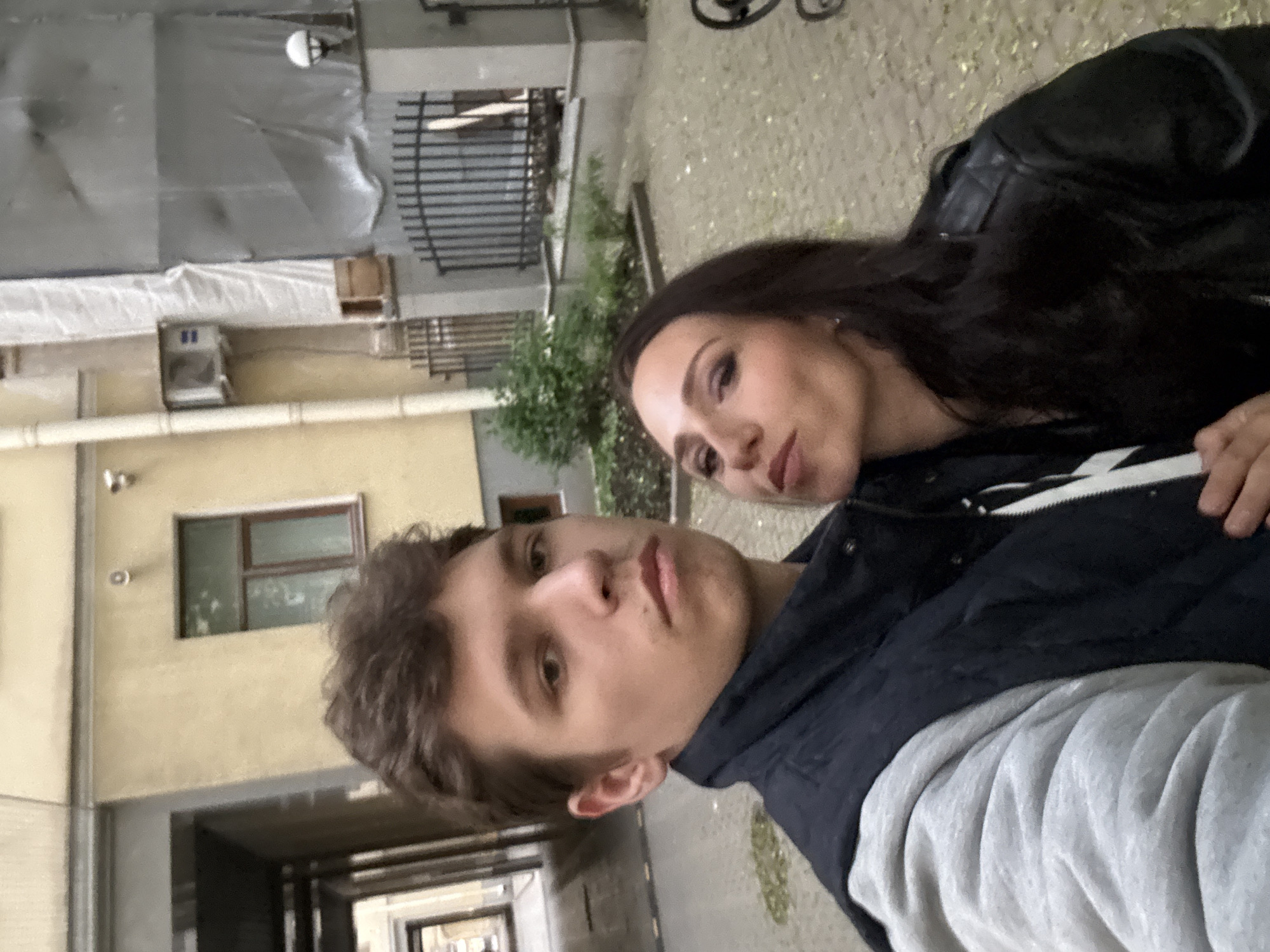 A selfie of two people, a man and a woman, standing close together in an outdoor urban setting that is consistent with the city of Saint Petersburg, Russia. The man is in the foreground, wearing a dark jacket over a grey sweatshirt. The woman is positioned behind him, partially obscured, with long, dark hair and wearing a black leather jacket. The background features a building with light-colored facade, a window, drain pipes, a fence, and some greenery. The setting is a courtyard or alleyway, characteristic of central Saint Petersburg. The light suggests overcast or late afternoon conditions.