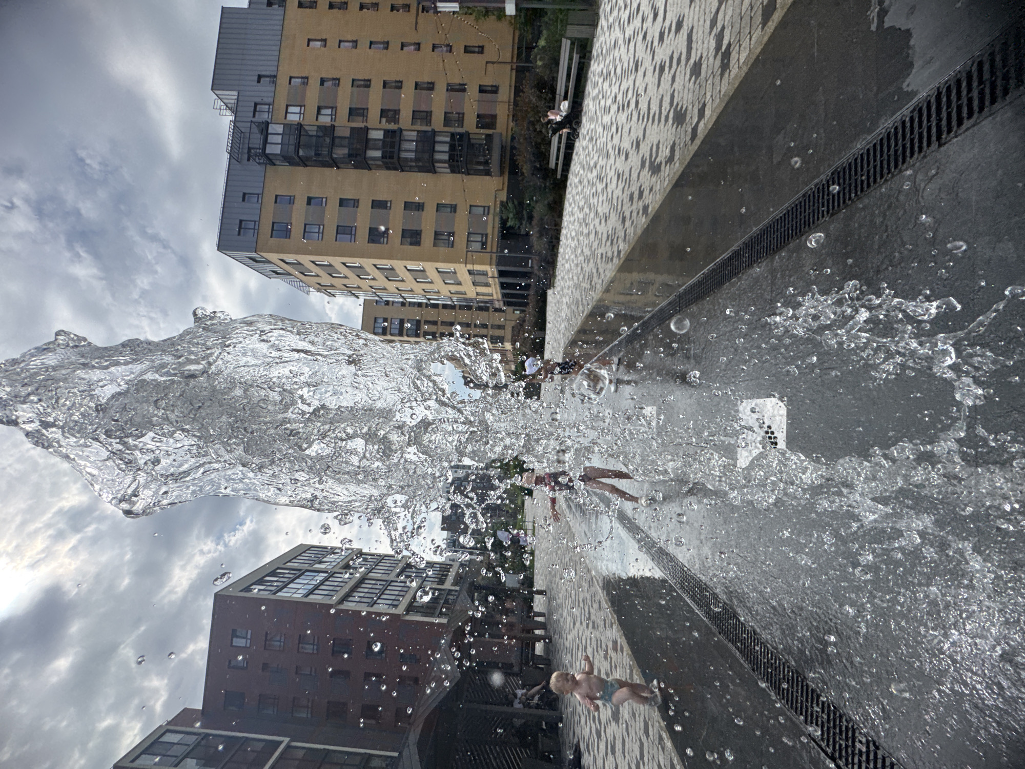 An outdoor shot captures a fountain's spray in the foreground, partially obscuring the background of a modern residential complex in Novoselye, Russia. The fountain's water is illuminated by sunlight, creating a dynamic, splash-filled effect. In the mid-ground, two children are visible playing in the water. To the left, a toddler in a diaper is running towards the fountain, while to the right, a young girl in a swimsuit and hat is standing with her arms outstretched, seemingly enjoying the water. The children are on a wet, dark paved area that appears to be part of the fountain's design, with a metal grate running alongside it. In the background, several multi-story buildings with yellow and brick facades dominate the scene. The sky is overcast with patches of sunlight breaking through the clouds. A few people are seated on a bench in the distance, and a stroller is nearby. The area is paved with a decorative stone pattern.