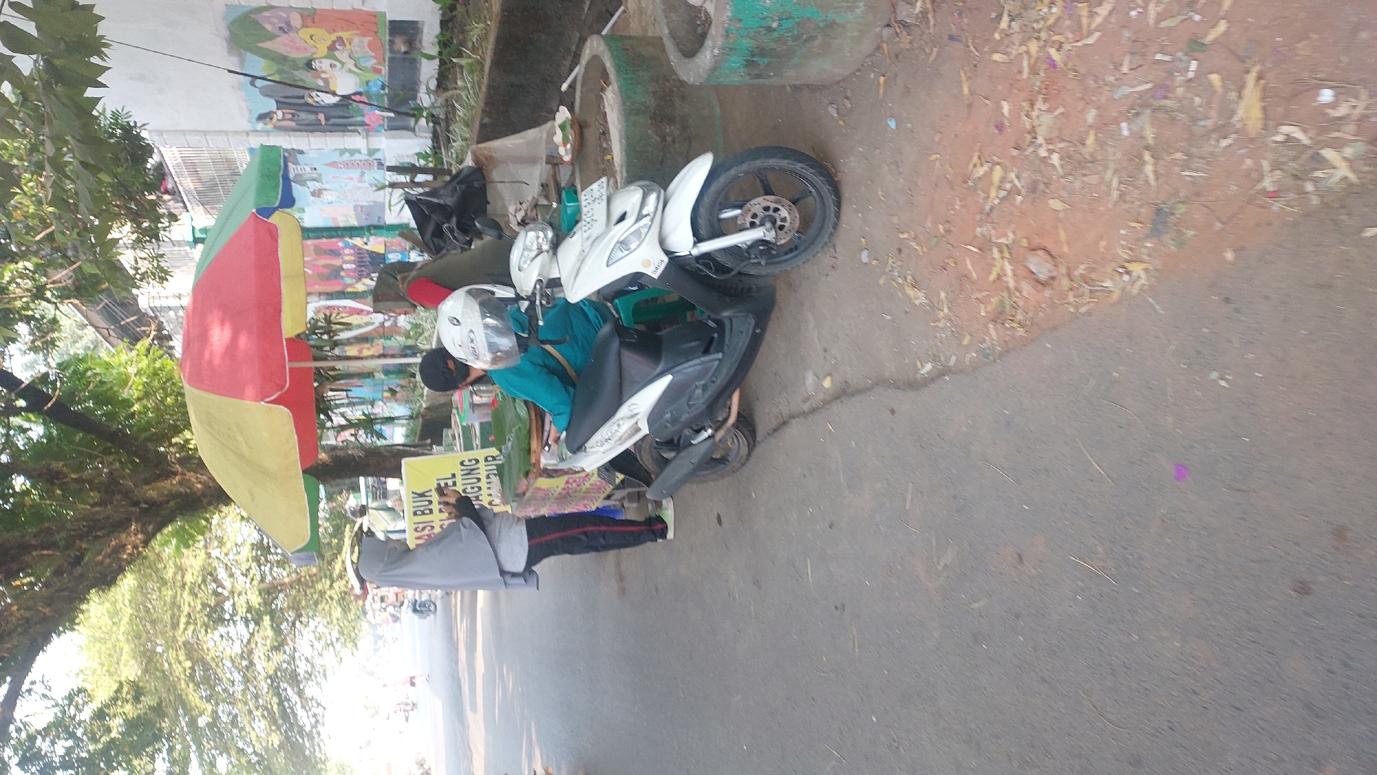 In Malang, Indonesia, a street scene unfolds beneath a colorful umbrella. A person wearing a grey hijab and black pants stands to the left, facing away from the camera. To their right, seated on a green stool, another individual in a teal shirt and white helmet is near a white scooter. Behind them, a food stall displays a sign with Indonesian text. The background features a wall adorned with murals and lush trees, suggesting an urban environment with natural elements. The ground is a mix of asphalt and dirt, littered with dry leaves, indicating an outdoor setting likely during daylight.