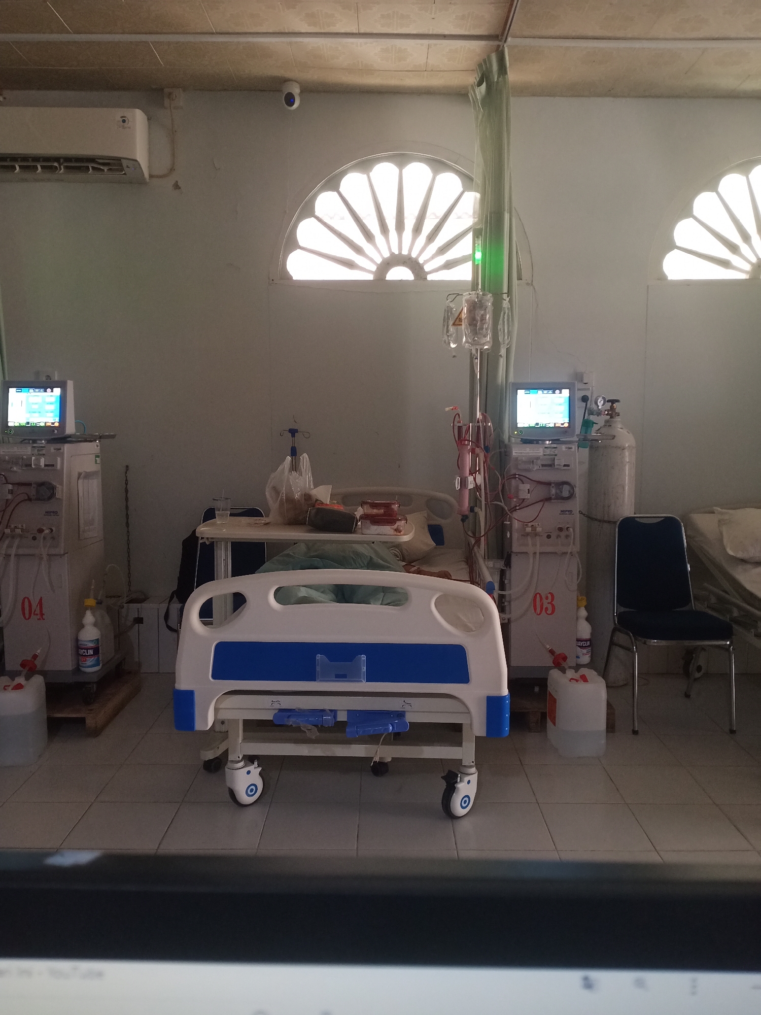 The image depicts a hospital room, likely a dialysis unit, located in Lhokseumawe, Indonesia. Two dialysis machines are positioned on either side of a hospital bed. The machine on the left is labeled with the number '04', and the machine on the right is labeled with '03'. A patient is partially visible in the hospital bed, lying down and covered with a sheet. An intravenous drip is suspended above the bed. Medical supplies and equipment are present, including bottles of liquid and a wheeled chair. The room features arched windows with decorative tracery, an air conditioning unit on the wall, and tiled flooring. The overall impression is one of a functional medical facility.