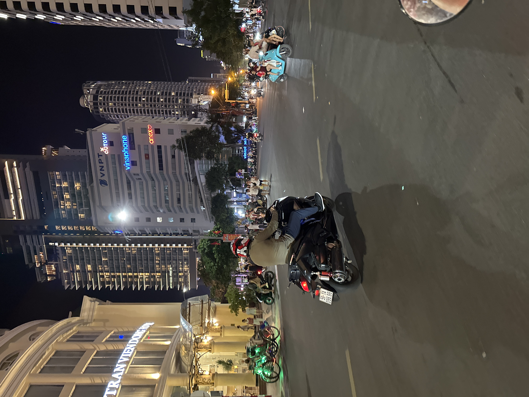 The image captures a nighttime street scene in Nha Trang, Vietnam, likely along a major thoroughfare given the presence of tall buildings and illuminated signage. The foreground is dominated by a dark-colored scooter with a rider wearing a red and white helmet and a khaki jacket. The scooter's license plate reads "59-YA 096.98." In the background, multiple scooters and a few pedestrians are visible, indicating moderate street activity. Several large, modern buildings with numerous lit windows tower over the scene, suggesting a commercial or residential district. Illuminated signs on these buildings include "VNPT," "autour," "vinaphone," and "anex♥." To the left, a building with a rounded facade and classical architectural elements is visible, featuring the illuminated text "TRAN VIEN DONG HOTEL." A distinct green, illuminated cyclopousse (pedicab) is parked on the sidewalk to the left. The street is paved and appears to be wet, reflecting some of the surrounding lights. The overall atmosphere is one of a bustling urban environment at night.