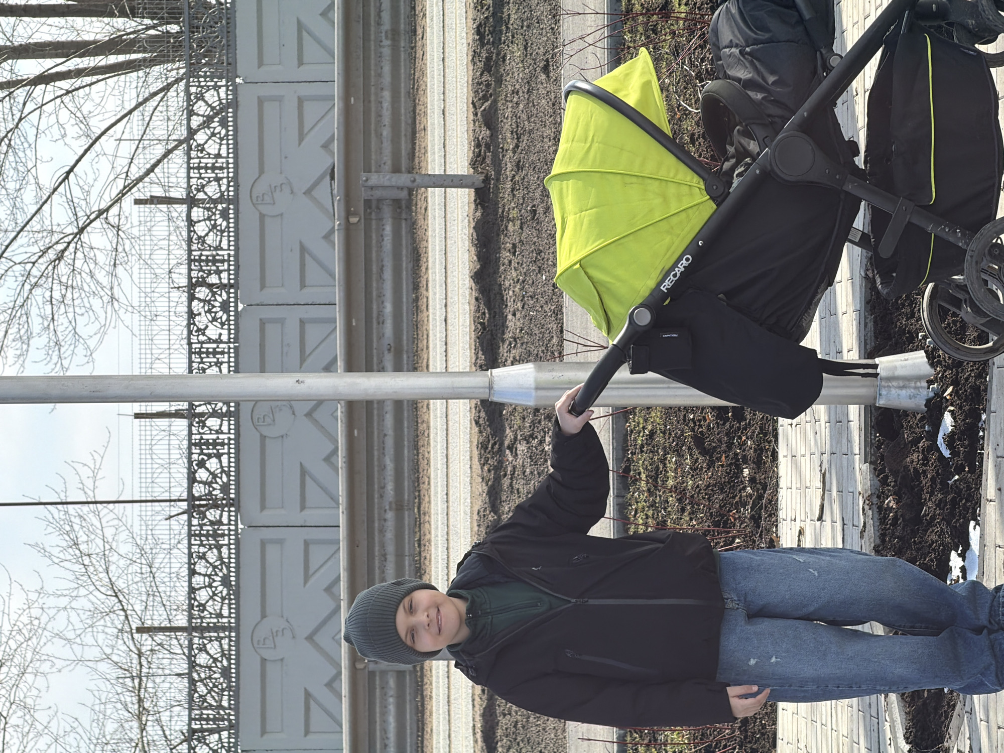 A boy in a dark jacket and gray beanie stands on a sidewalk next to a Recaro stroller with a chartreuse green hood. The stroller rests on a paved surface with patches of dirt. A metal guardrail runs parallel to the sidewalk, and behind it is a fence with decorative metalwork. Bare trees are visible against a pale sky. The location is likely Votkinsk, Russia, based on the decorative metalwork on the fence which is a landmark of the city.