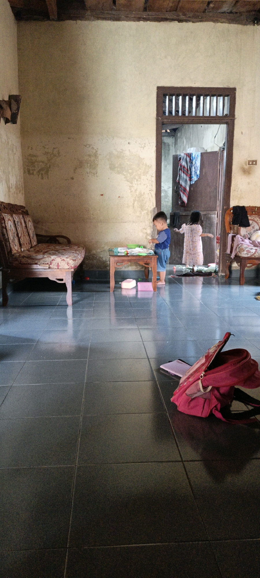 The image depicts an indoor scene, possibly a living room or common area, within a dwelling in Bunut, Indonesia. The room has a rustic and lived-in aesthetic, with textured, off-white walls showing signs of age and wear. The ceiling features exposed wooden beams, suggesting traditional construction. Dark grey tiled flooring covers the ground, reflecting light and the objects within the room.
In the mid-ground, a young boy, wearing a blue and white striped shirt and denim shorts, stands at a small, low wooden table. He appears to be engaged with items on the table, possibly toys or school supplies. To his right, a doorway leads to another area of the house, where a young girl in a light-colored dress is partially visible, facing away from the camera. Clothes are draped over the doorway and within the adjacent room.
To the left of the frame, a three-seater sofa with patterned upholstery sits against the wall. The sofa appears to be made of wood with ornate carvings. On the floor in the foreground, a bright red backpack lies open, with a notebook or flat object tucked inside. Other objects, including what appear to be plastic containers, are scattered on the floor near the table. The overall impression is of a domestic environment with children present, reflecting a casual, everyday setting.
