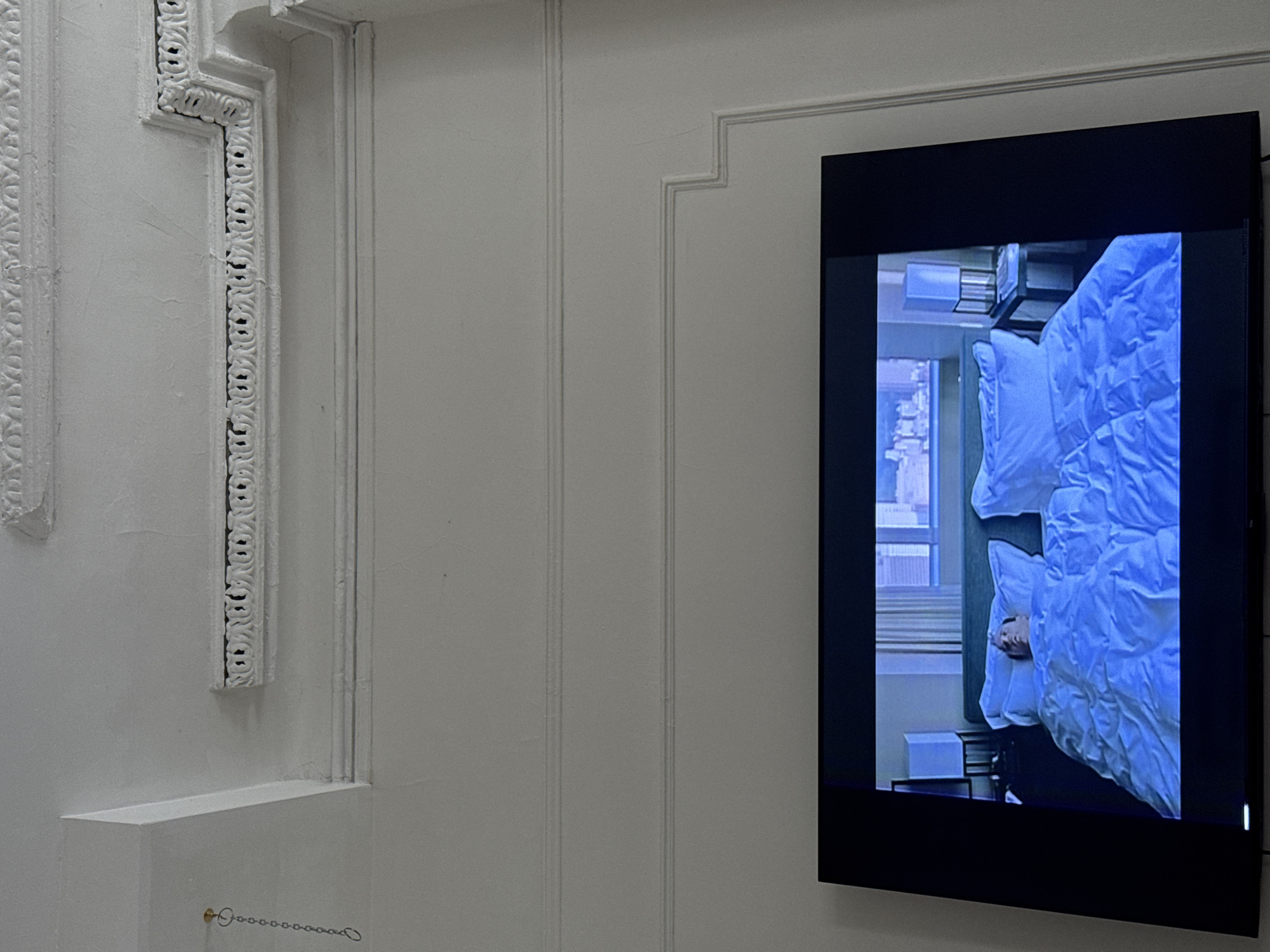 A white wall with decorative trim features a modern black-framed screen displaying a video of a bed in a hotel room. The video captures the act of a person lying in the bed, with a window visible in the background indicating an urban setting, possibly in New York City, United States of America.