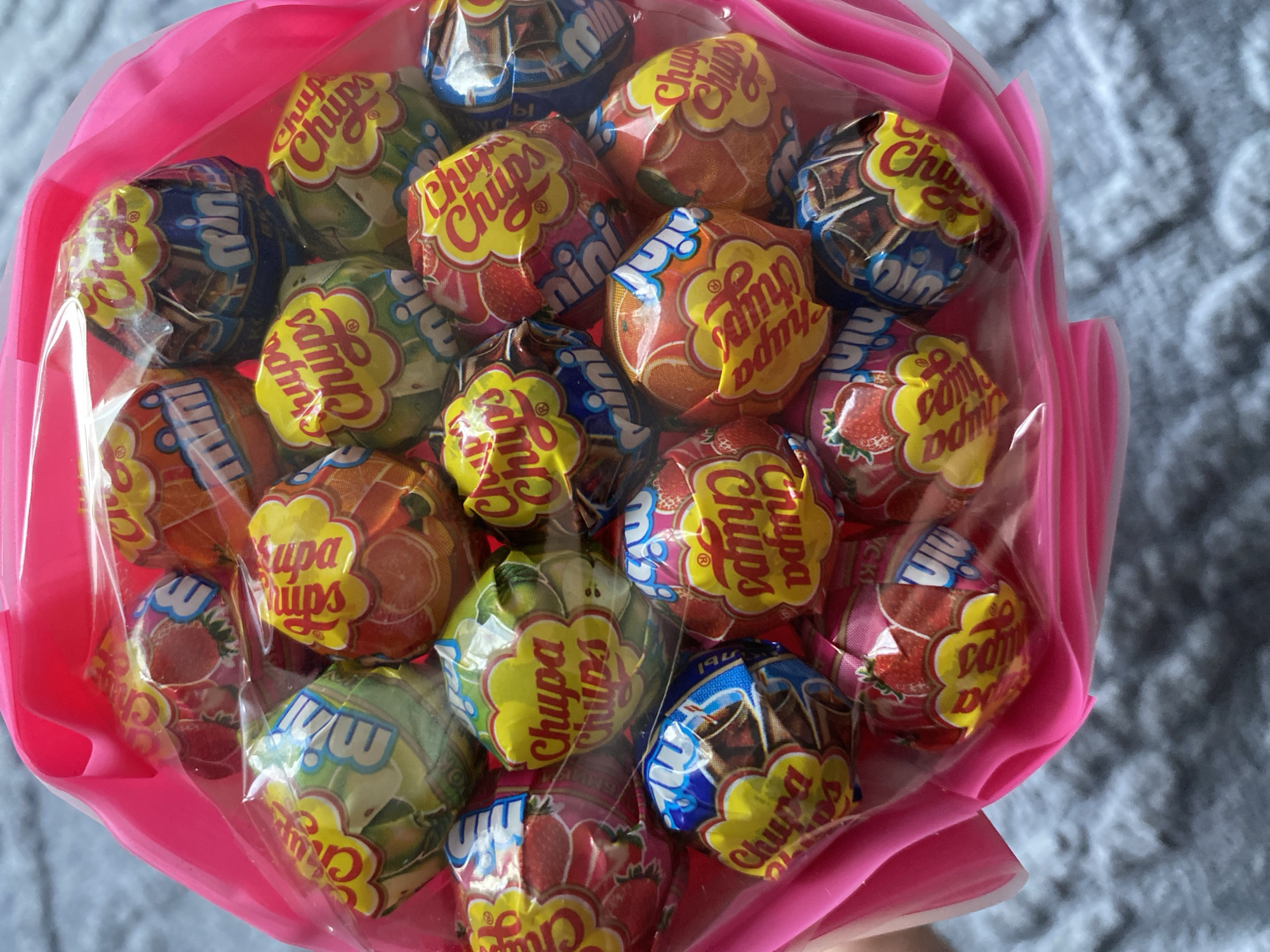The image displays a vibrant close-up of a "bouquet" composed entirely of Chupa Chups lollipops. Numerous lollipops, each individually wrapped, are densely packed together, forming a colorful arrangement. The wrappers showcase a variety of flavors, including prominent designs featuring strawberries, oranges, and apples, alongside other patterns in blues, browns, and greens, suggesting flavors like chocolate or cola. The iconic yellow daisy-shaped "Chupa Chups" logo in red text is clearly visible on most of the wrappers, with some also featuring the word "Mini."
The collection of lollipops is enclosed in a transparent plastic film, which is then gathered at the base and further wrapped in bright pink plastic or cellophane, mimicking the presentation of a flower bouquet. The background is softly blurred, showing a light grey, textured fabric, possibly a blanket or rug, indicating an indoor setting. No individuals are visible in the frame, and the time of day or specific weather conditions are not discernible. The arrangement suggests a celebratory gift or a special treat. While the provided location context is Noviy, Russia, no specific visual cues within the image confirm this location.