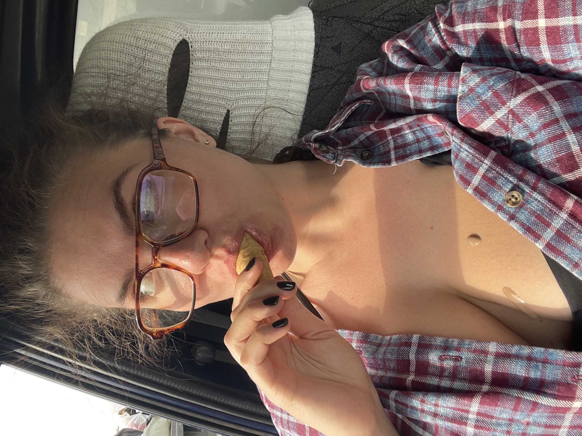 A woman with brown hair and glasses is lying down, possibly in a vehicle, and eating a potato chip. She is wearing a maroon and blue plaid shirt over a black top. A grey knitted balaclava is next to her head. The image does not provide enough information to determine the location in Molokovo, Russia.