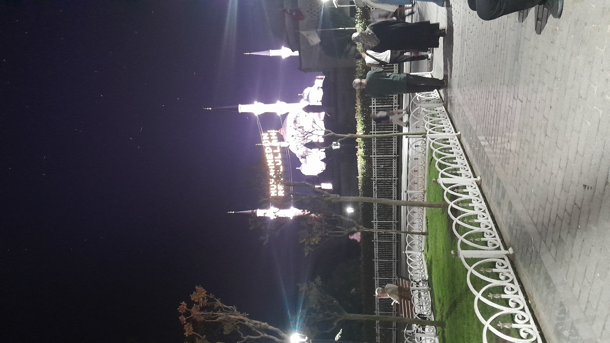 This is a nighttime photograph of the Blue Mosque (Sultan Ahmed Mosque) in Istanbul, Turkey. The mosque is illuminated, and a banner with Arabic script, likely a religious message, is visible across the front of the building. Several minarets and domes of the mosque are prominent in the background.
In the foreground, a paved walkway is visible, with people walking and standing. To the left of the walkway is a section of manicured lawn enclosed by a decorative white metal fence. A person is seated on a bench within this fenced area, facing away from the camera and appearing to be looking at a device. Small trees with bare branches are planted within the fenced area and along the walkway.
The sky is dark, indicating it is nighttime, and a few faint stars are visible. The overall lighting suggests an urban environment at night, with artificial illumination from the mosque and nearby light sources. The scene conveys a sense of public space and people visiting a significant cultural and religious landmark.