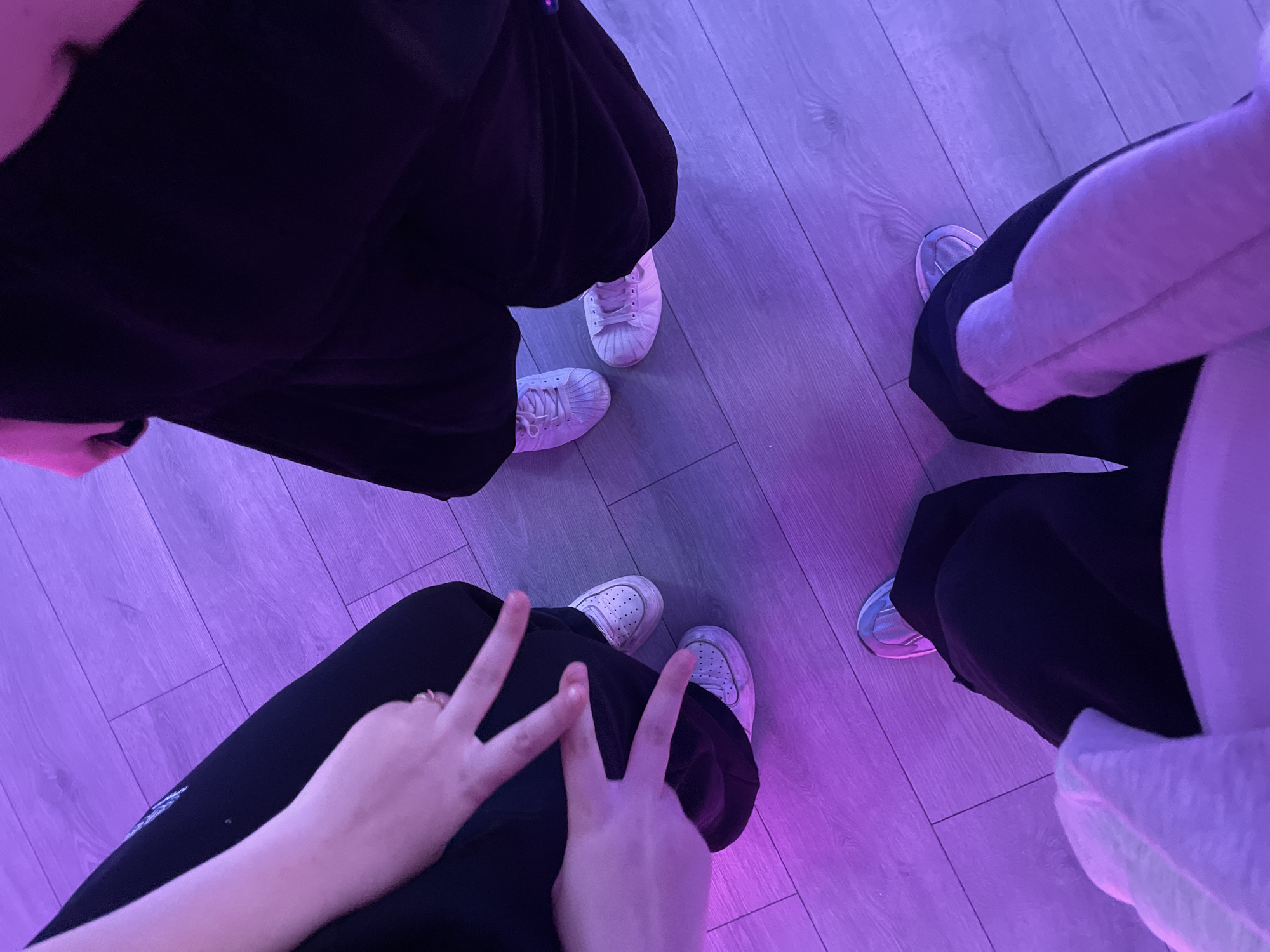 An overhead shot captures the lower bodies of three individuals, possibly in Saint Petersburg, Russia, standing or sitting on a light-colored wooden or laminate floor bathed in vibrant purple artificial light.
In the upper left portion of the frame, one person wears loose-fitting black pants and white sneakers with textured toe caps. Their feet are positioned slightly apart. To the lower center, another individual is seen wearing black pants and white perforated sneakers. This person's hands are prominent, with both performing a "peace sign" gesture, suggesting a playful or casual mood. On the right side of the image, the third person wears black pants paired with light-colored sneakers, possibly white or light blue, and a light-colored (likely white or grey) hoodie or jacket, part of which drapes over their legs.
The strong, consistent purple illumination across the entire scene suggests an indoor setting with specialized lighting, such as a studio, club, or a room equipped with LED lights. No specific time of day is discernible due to the artificial light, but it hints at an evening or night-time activity. There is no visible text in the image. The overall impression is a casual, social gathering, possibly a photoshoot or a relaxed moment among friends.